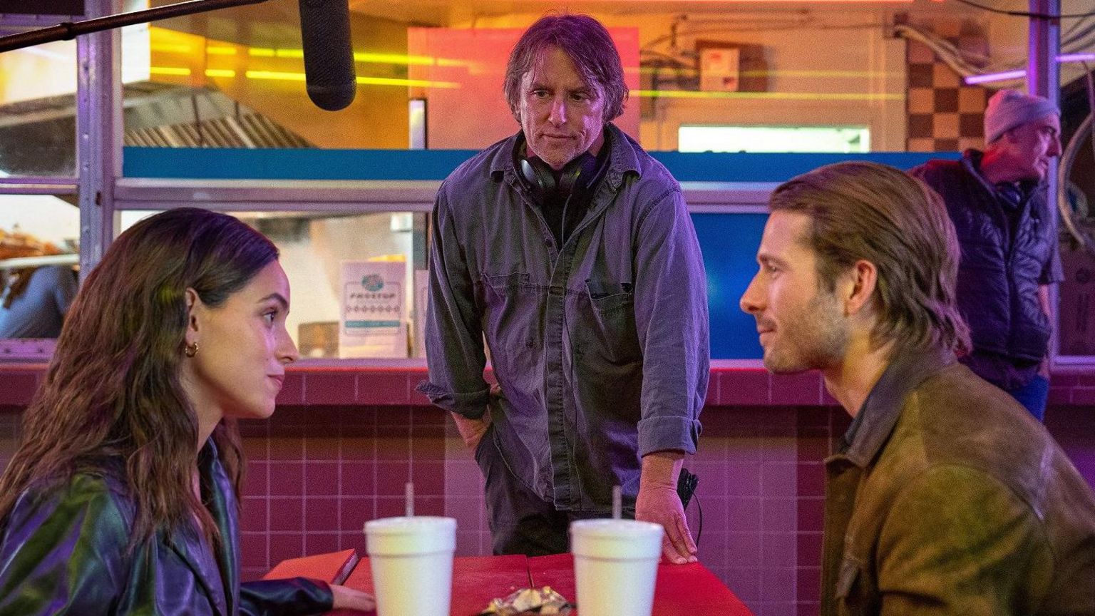 Director Richard Linklater watching Adria Arjona and Glen Powell filming a scene playfully looking at each other across a bench at a diner