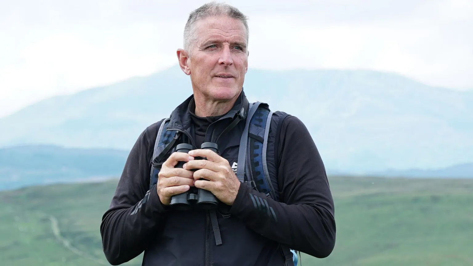 S4C Iolo Williams stands with stunning green scenery behind him. He wears a black jacket and a navy rucksack. He hols binoculars in his hands which are by his chest.