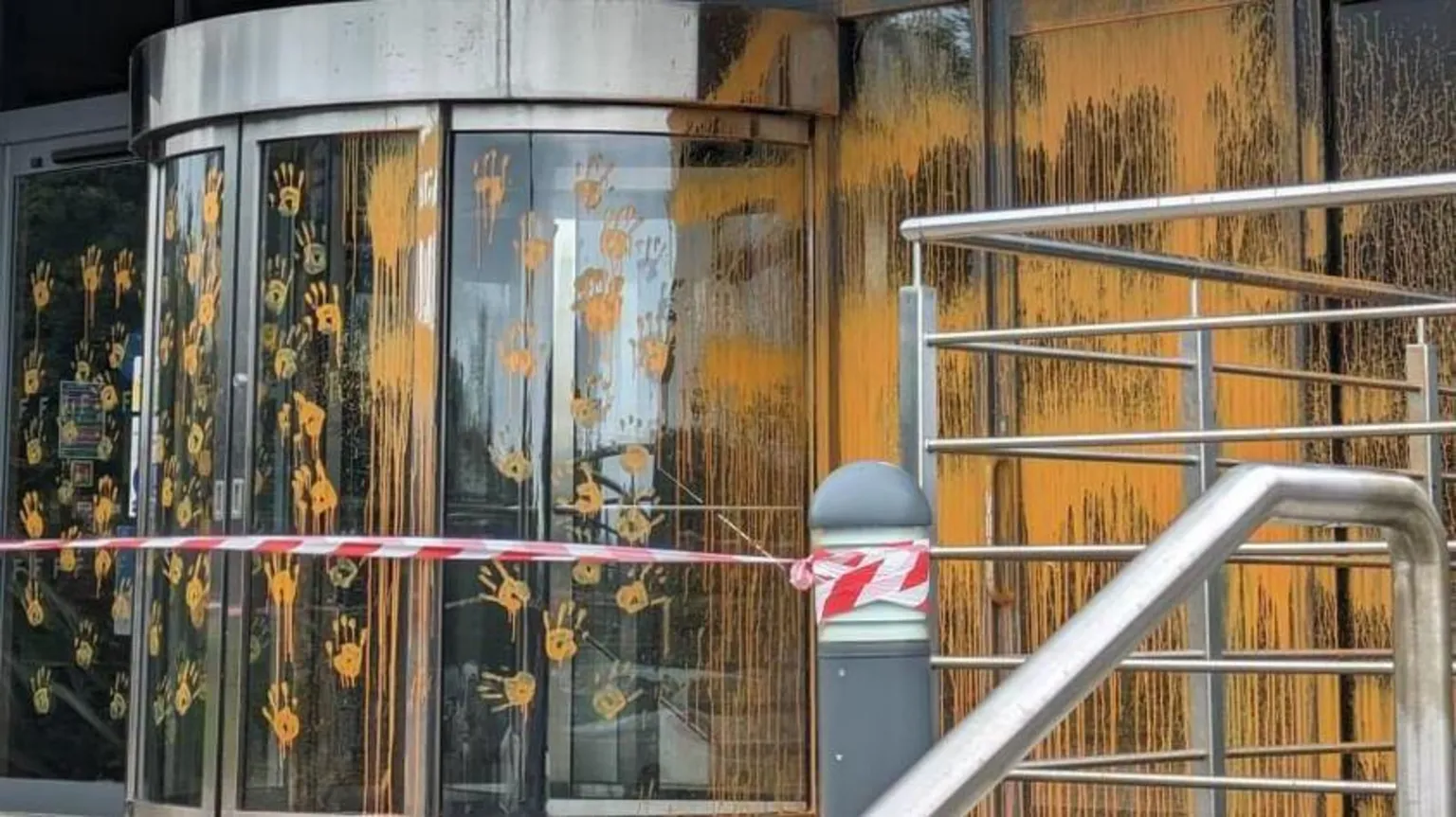 The image shows a modern glass building entrance covered in orange paint and yellow handprints. Red and white police tape cordons off the area. Metal railings and steps are visible, with paint dripping down the glass surfaces of what appears to be a university building entrance.