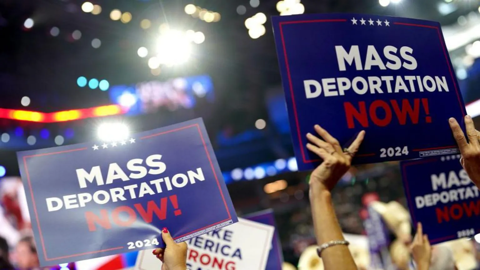 A close-up, low-angle shot of supporters at a political convention holding signs high above their heads. The prominent blue signs display the message &ldquo;MASS DEPORTATION NOW!&rdquo; in large white and red font. The background is filled with blurred arena lights and other campaign signage, including a partially visible &ldquo;MAKE AMERICA STRONG&rdquo; sign.