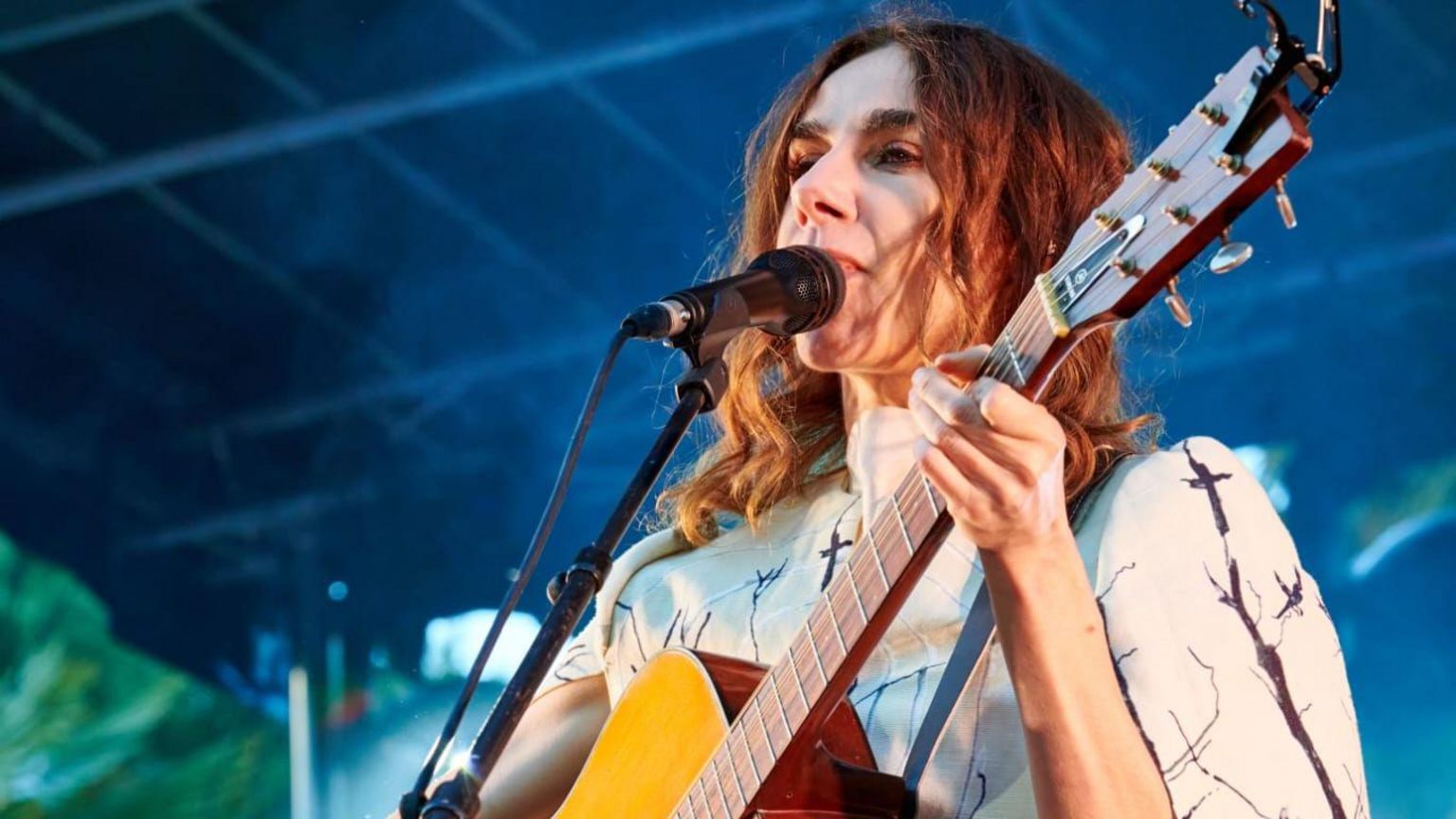 PJ Harvey holding an acoustic guitar and singing into a microphone. She has shoulder length curly brown hair and is wearing a fitted white dress.