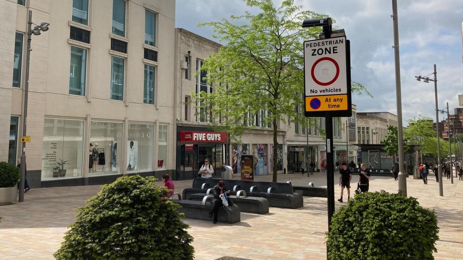 A street sign with 'Pedestrian Zone' stands in front of a shopping high street with a handful of pedestrians