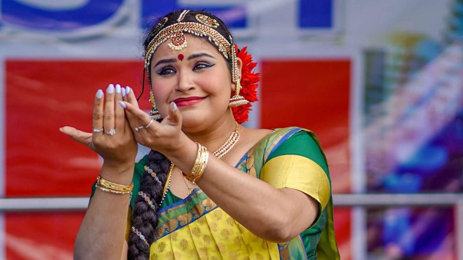 A woman with her hair in a long plait, wearing a green and yellow sari and a gold beaded headpiece, holding her arms outstretched in a dance