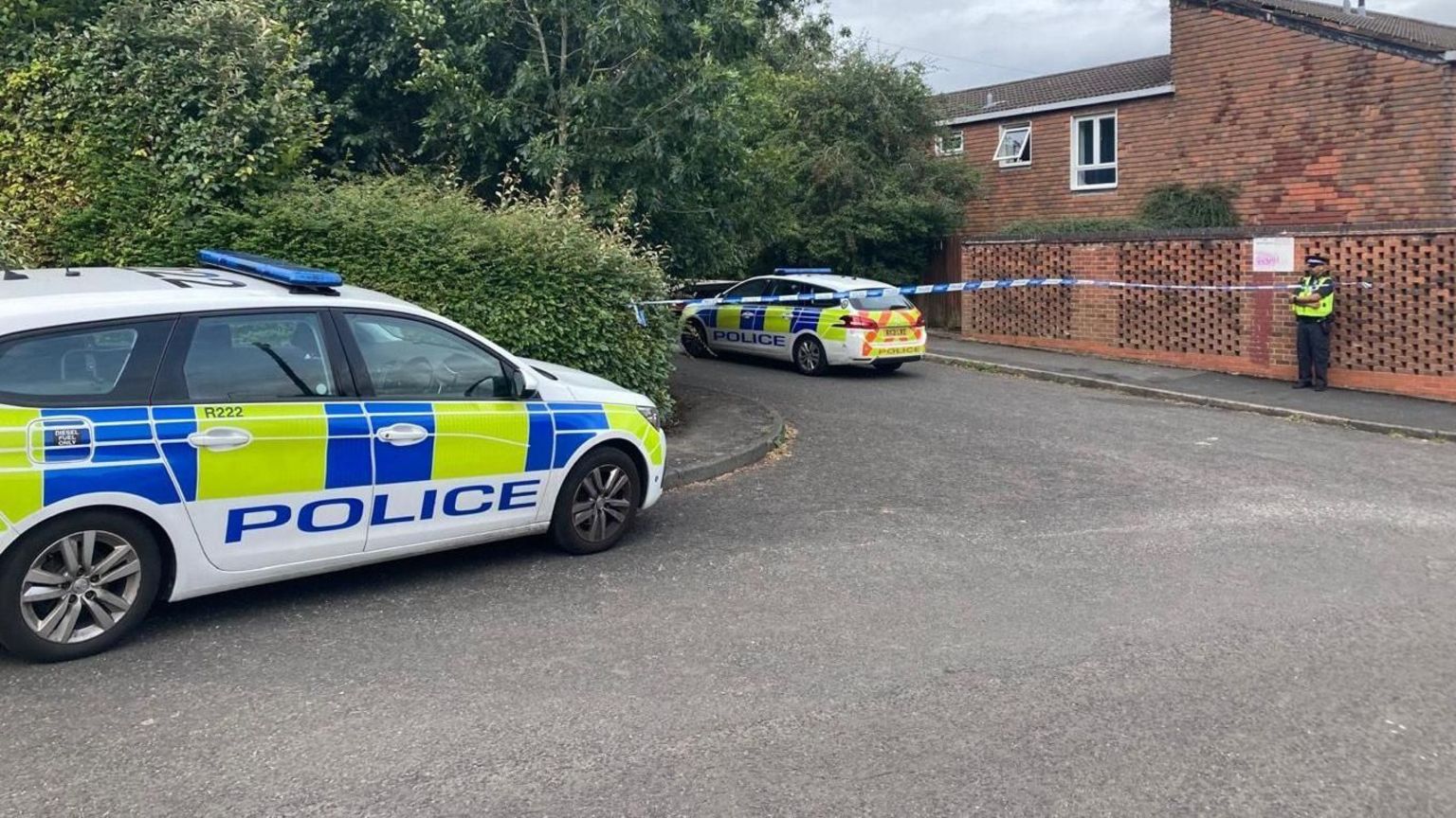 Two cars, a police cordon and an officer stood by a wall