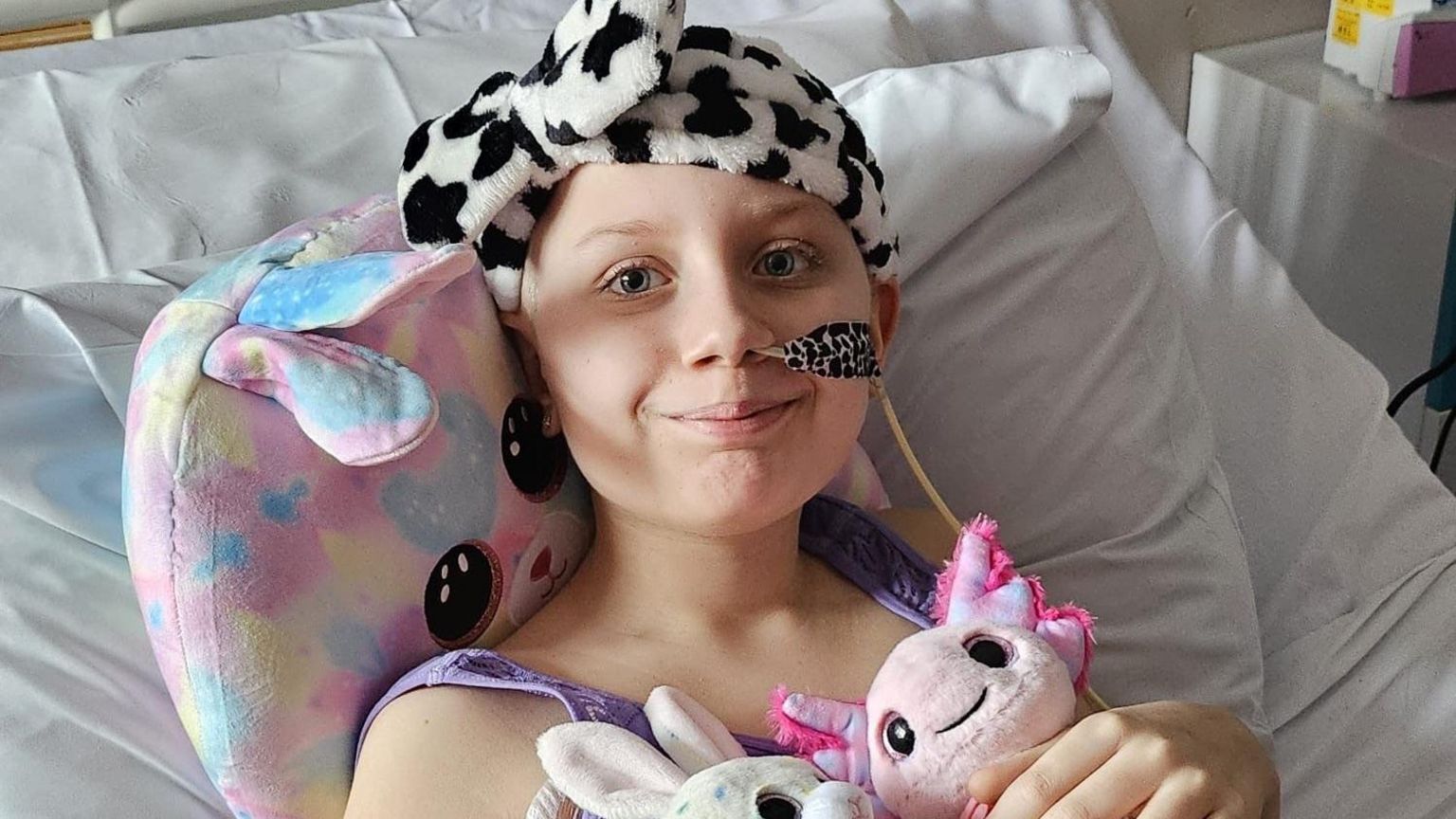 Megan. She is lying in a hospital bed with three cuddly toys. She has a tube in her nose and a black and white fluffy headband on. She is smiling at the camera.  