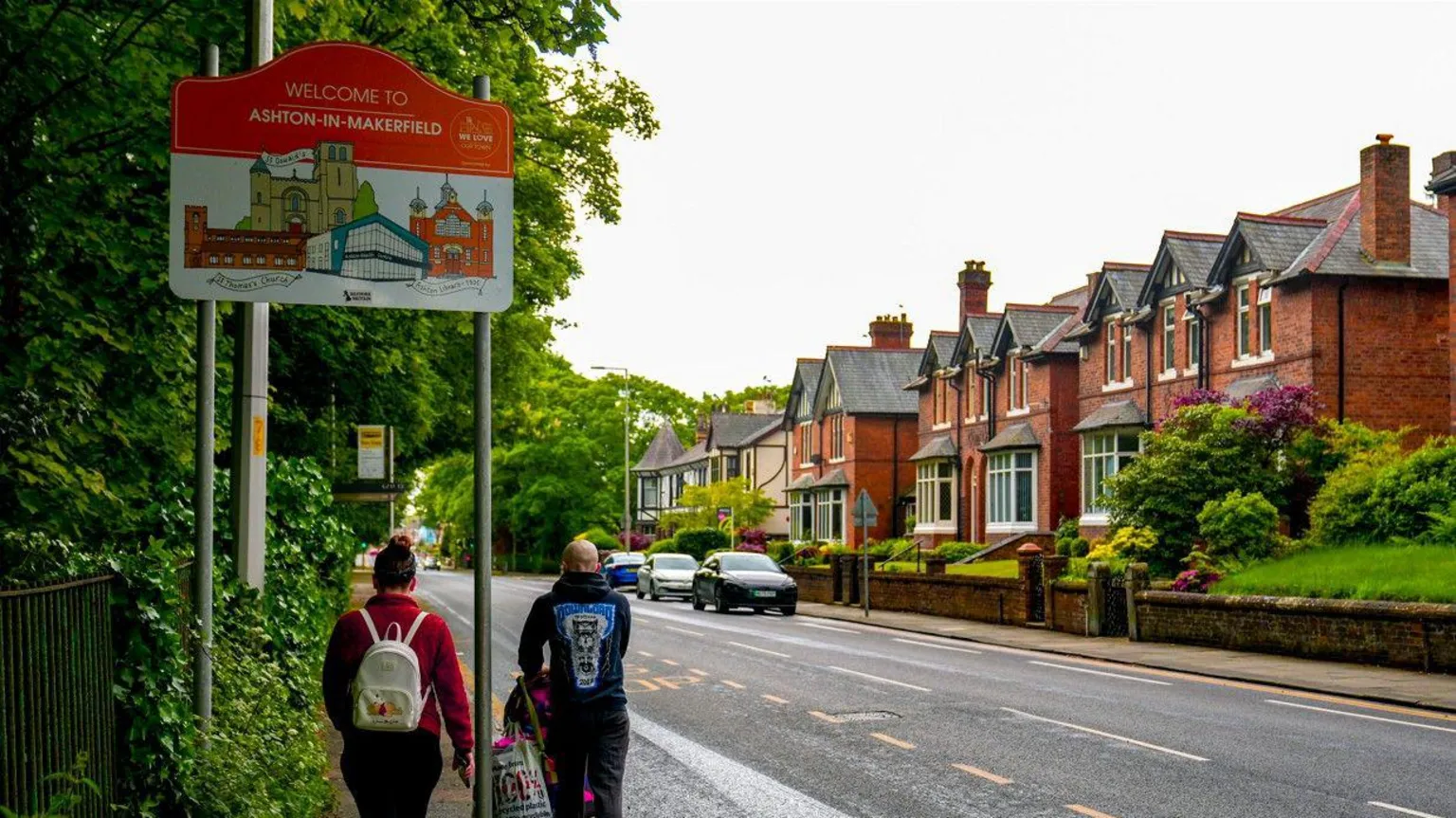 PA Media Two people walk under a sign, one pushing a pram, along a roadside in Ashton-in-Makerfield, Greater Manchester.