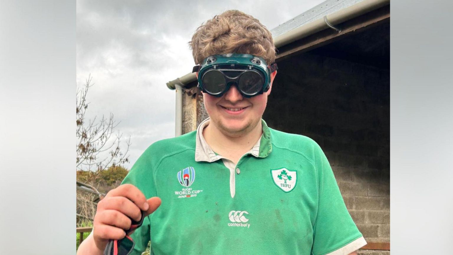 Ryan Taggart farmer pictured wearing a pair of safety goggles in front of a farm building. He is wearing a green Ireland rugby jersey which has dirt stains on it. Ryan has a pair of sunglasses in his hand which is raised. 