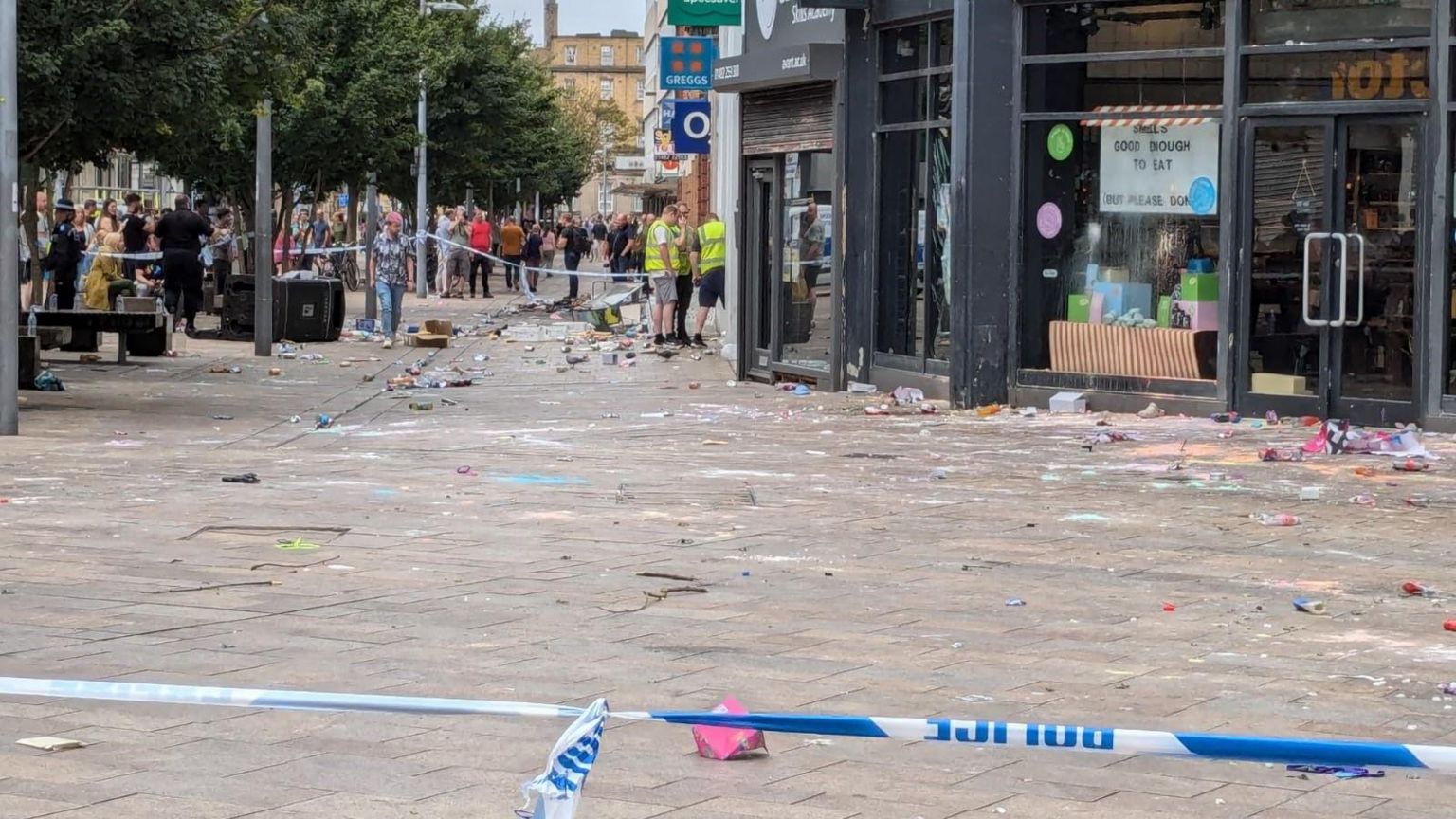 A police cordon outside shops in the city centre with windows smashed and pieces of glass on the floor. Members of the public look on and workers in high-vis vests are looking at the damage