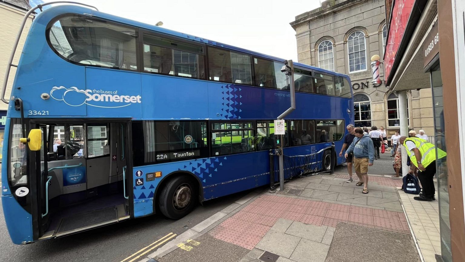 Blue double decker seen from the right, with a pedestrian guardrail pressed against its left-hand side as people watch on