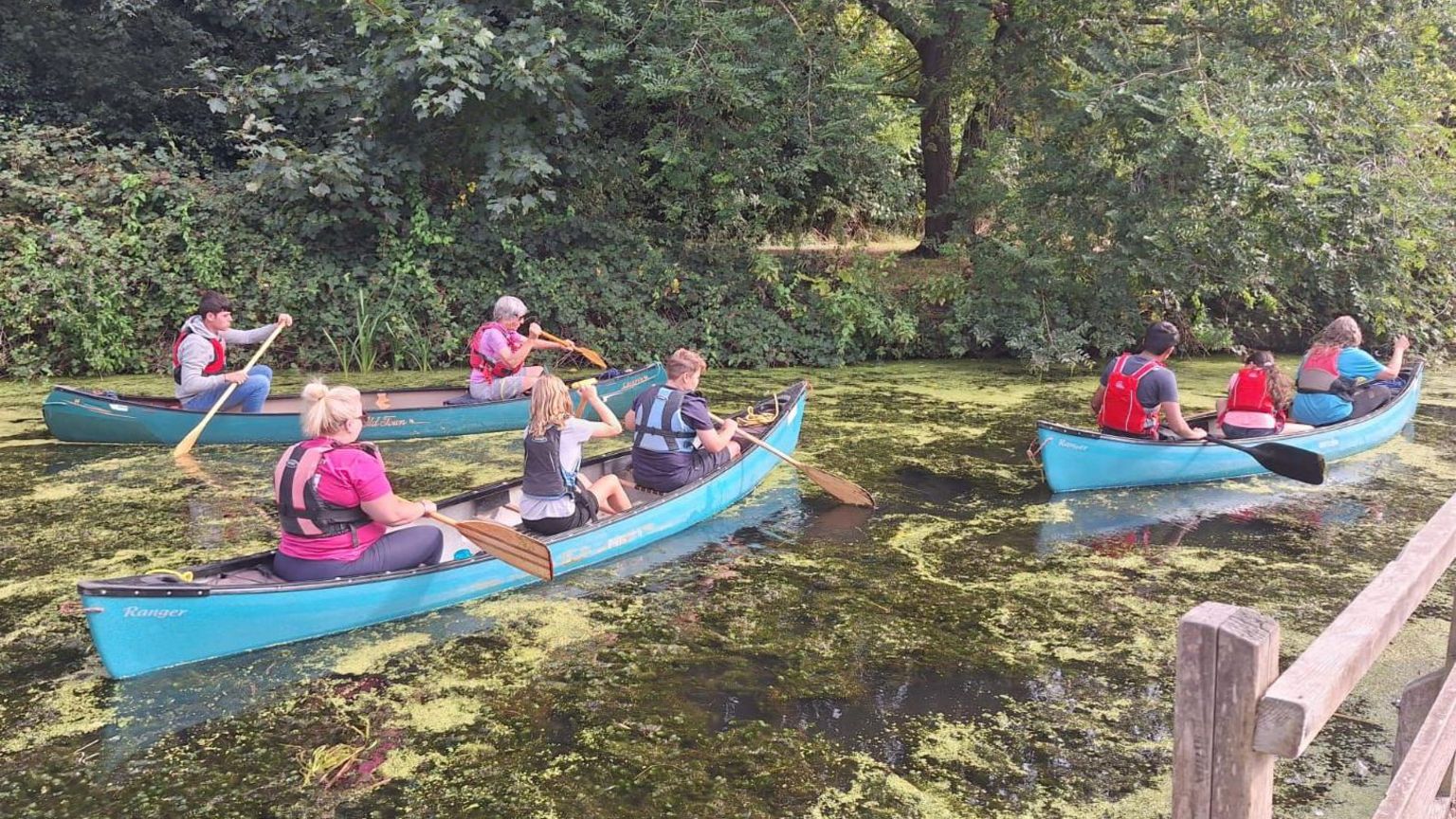 Eight people in three turquoise canoes paddle along a river with a fence in the foreground and trees in the background. There are three people in the first canoe, three in the second and two in the third. All are wearing lifejackets.