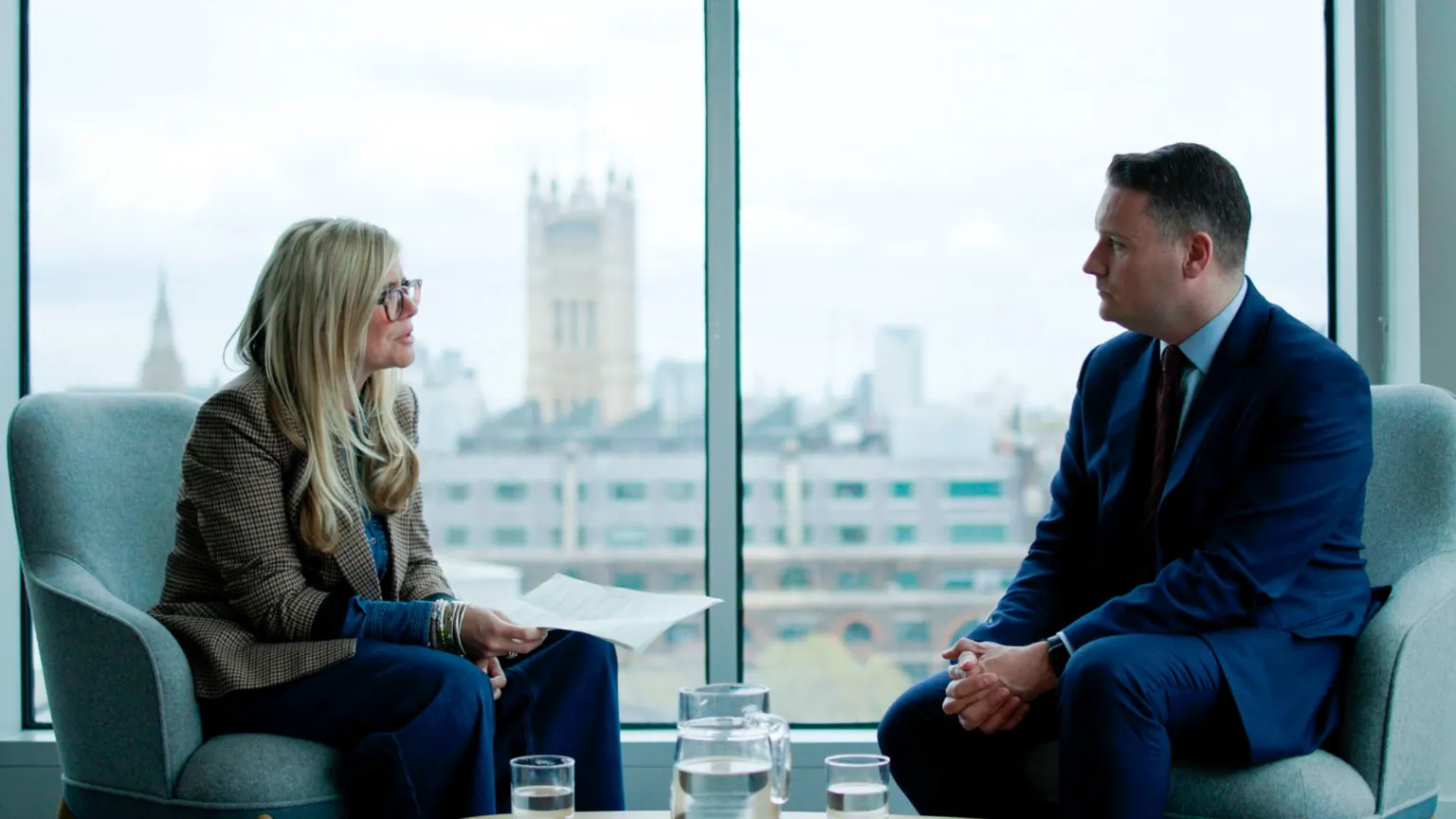 The BBC's Emma Barnett sits on the left in an armchair holding some papers while to the right is former Health Secretary Wes Streeting sitting opposite. He has short brown hair and wears a navy suit, a blue shirt and a dark tie. He clasps his hands together, while behind them Parliament's Victoria Tower can be seen through the window, rising up over the rooftops.
