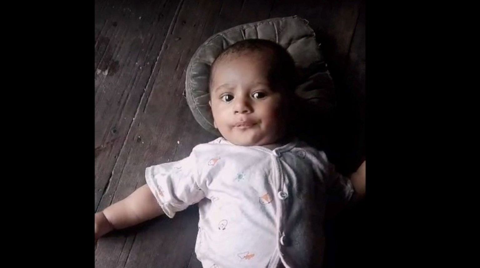 Fayaz's son looks at the camera. The baby is lying on a wooden surface with a pillow behind his head, wearing a baby grow. 