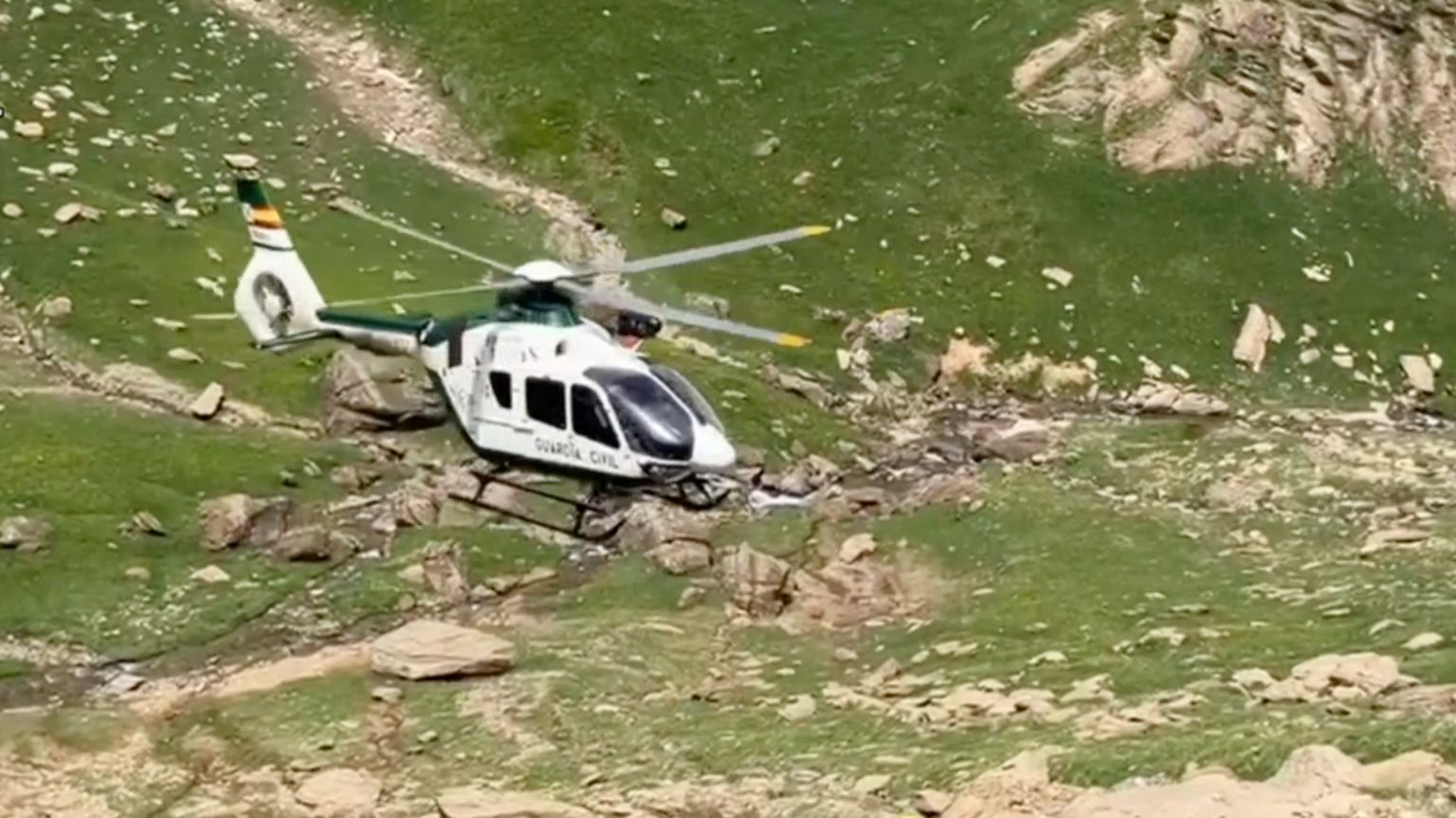 Light sandy coloured rocks on the grassy mountainous landscape of the Pyrenees with a white helicopter flying above the ground bearing the insignia Guardia Civil.