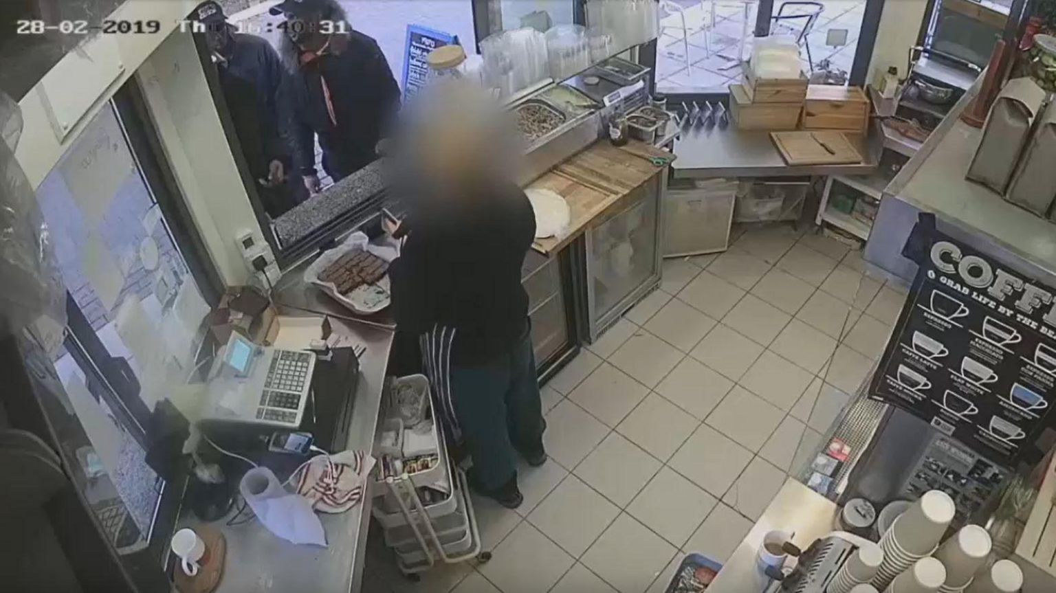 CCTV image of two men at the counter of a kitchen shop, man behind the count's face is blurred out