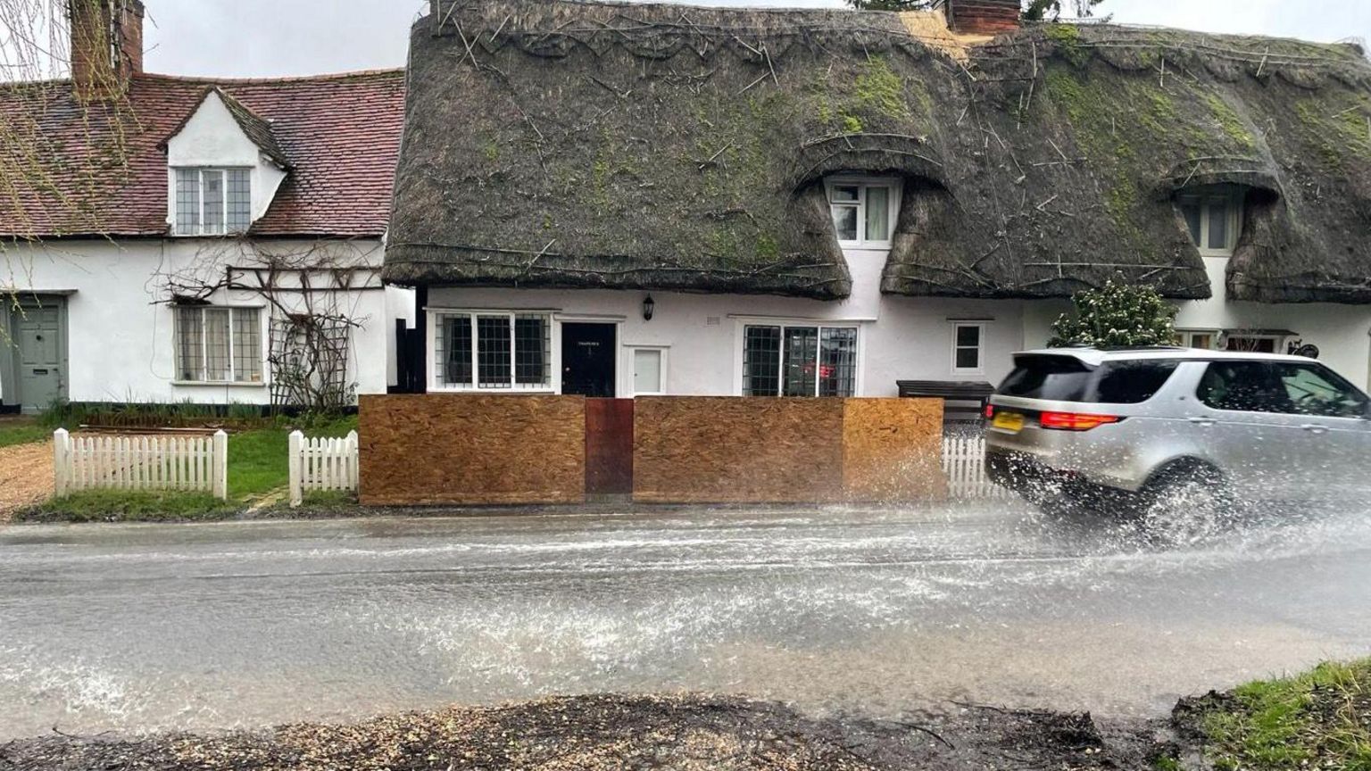 Cottage with wooden board barricading around it and a car driving past in flood water