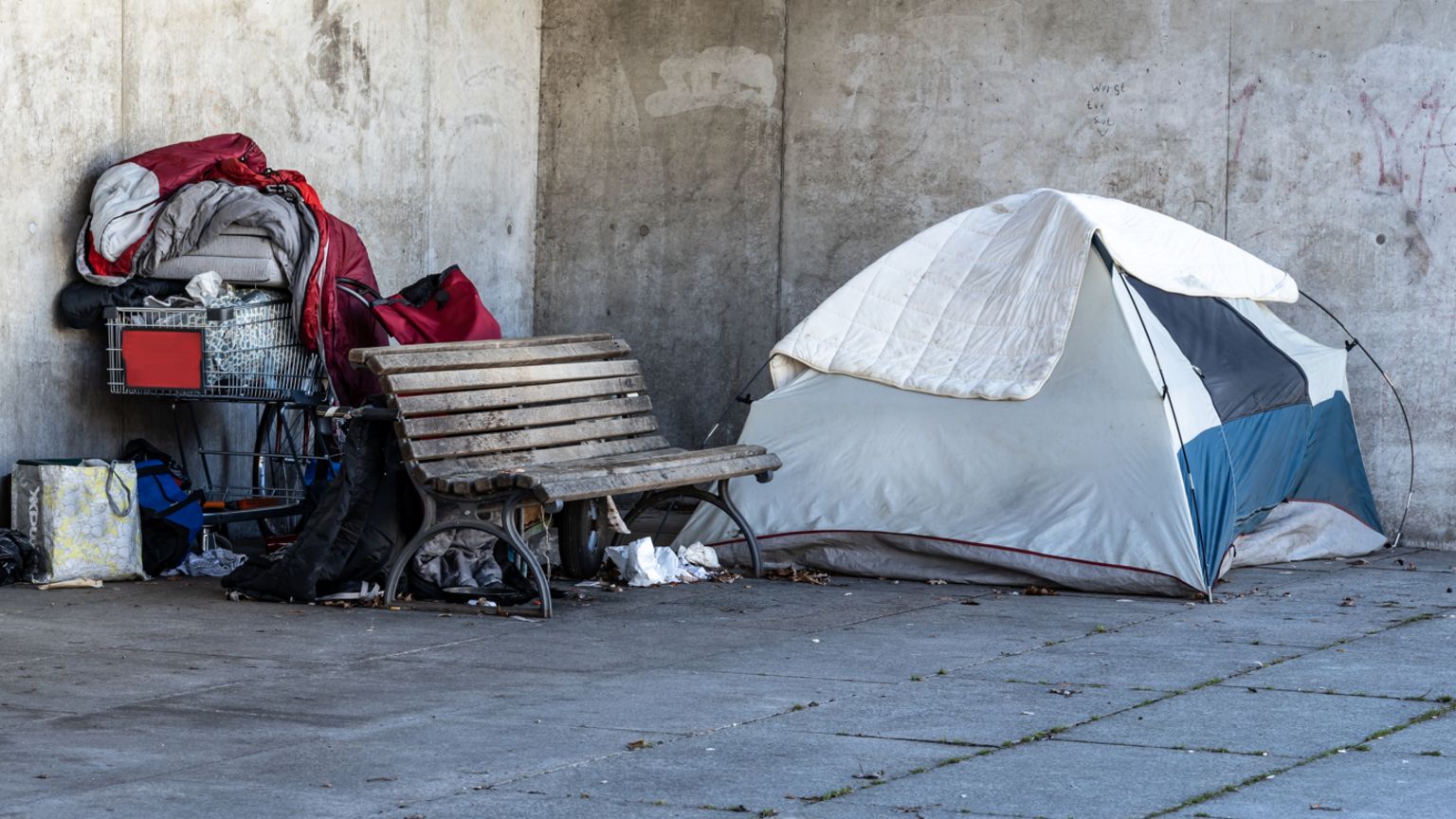 A tent and the belongings of a homeless person next to a bench outside