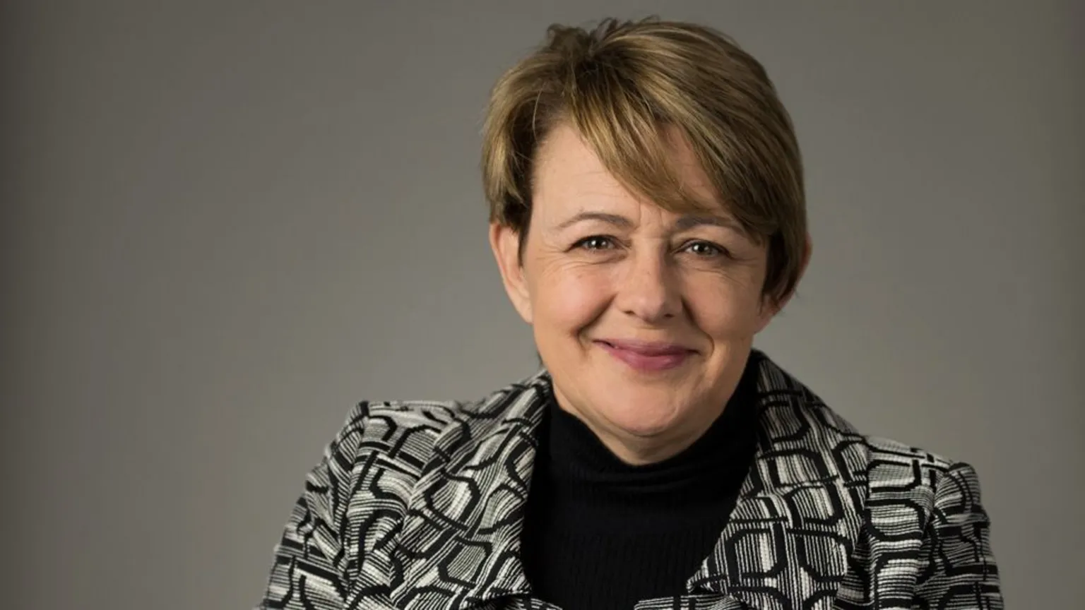 Portrait of a white woman with short ashy brown hair smiling at the camera, wearing a black and white blazer and a black shirt. 