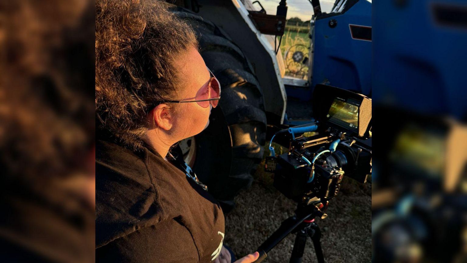 Kathryn Gadd wearing sunglasses with her hair tied back. She is looking at the screen of a camera in front of her, which is sitting on a tripod.