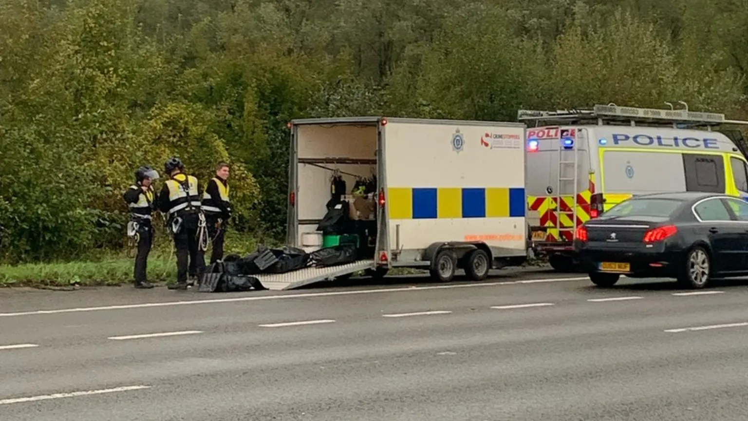 The image shows police officers in high-visibility vests standing beside a police trailer and van on a road. The officers appear to be handling equipment near the open trailer. A dark car is visible, and the scene is set against a backdrop of green vegetation along the roadside.