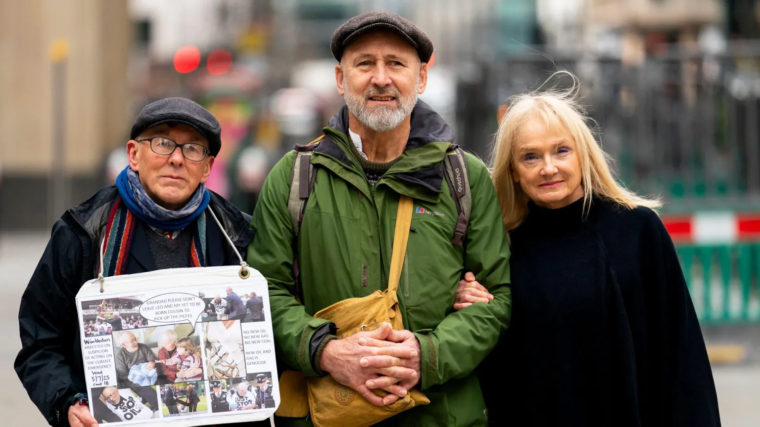 Three people stand on a city street. The man on the left wears glasses, a cap, and blue scarf while holding a white poster with photos and text. The middle man wears a green jacket and cap. The blonde woman on the right wears black clothing.