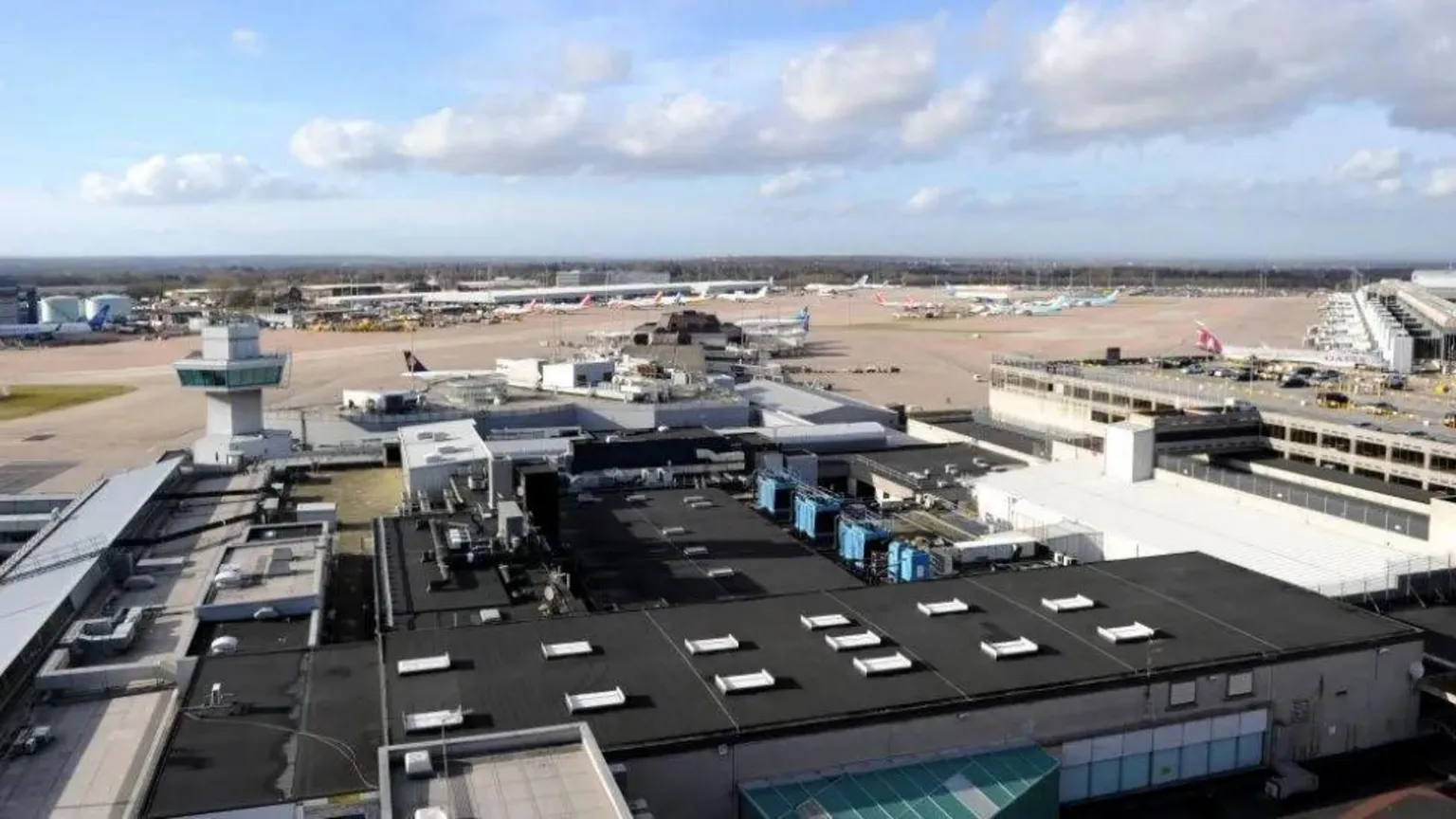 This aerial view shows an airport terminal with a large flat roof featuring multiple white ventilation units. The terminal building has glass facades and connects to jet bridges. In the background, aircraft can be seen parked on the tarmac, with runways and airport infrastructure extending across the landscape under a cloudy sky.