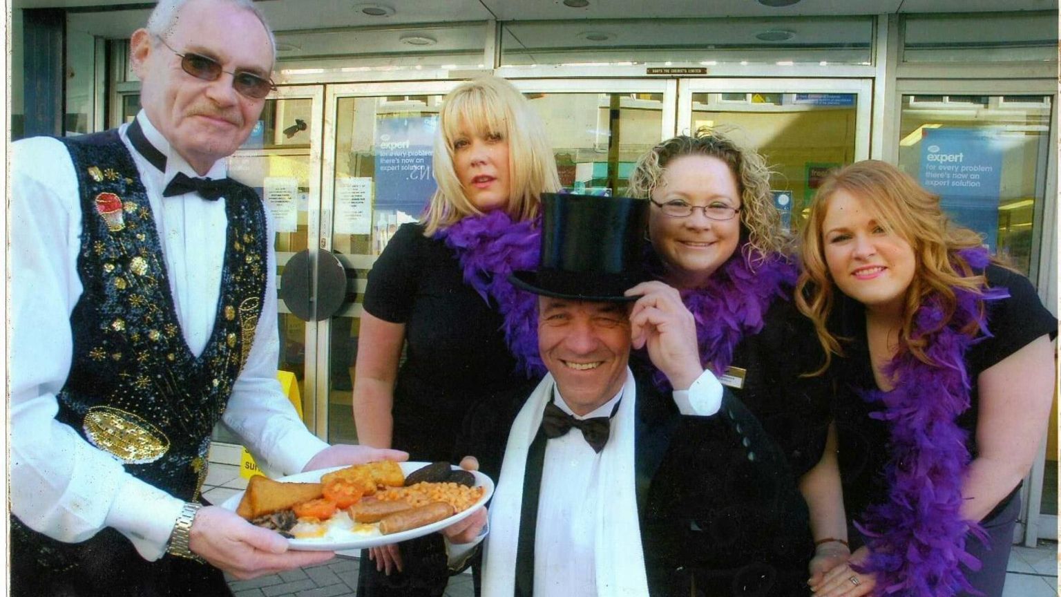 Mr Walker in top hat and tails being served breakfast on his Big Issue pitch outside Boots in Weston-super-Mare surrounded by ladies in feather boas and a man in bow tie and waistcoat