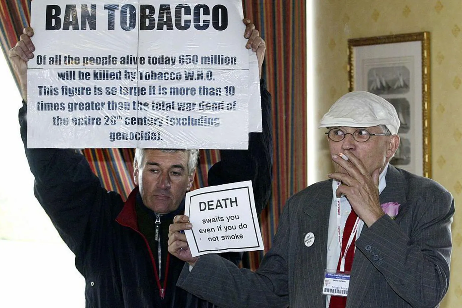 Getty Images David Hockney and an anti-smoking protester in 2005. The protester is holding up a sign calling for tobacco to be banned.  Hockney is holding up a small counter sign saying that people die even if they don't smoke.  He is smoking.