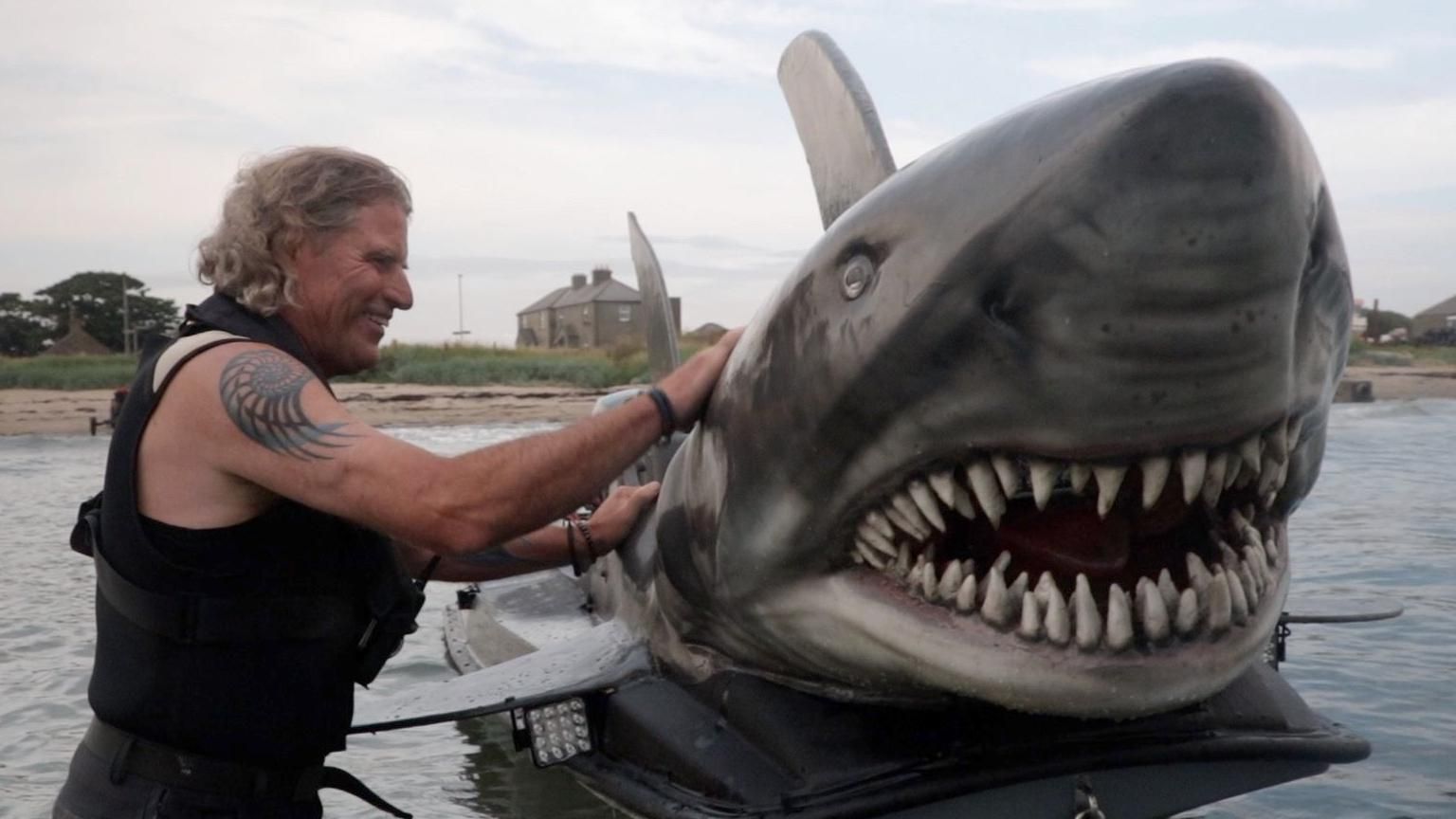 Fin Bowron in the ocean with his shark boat, he stands beside it and pets the head