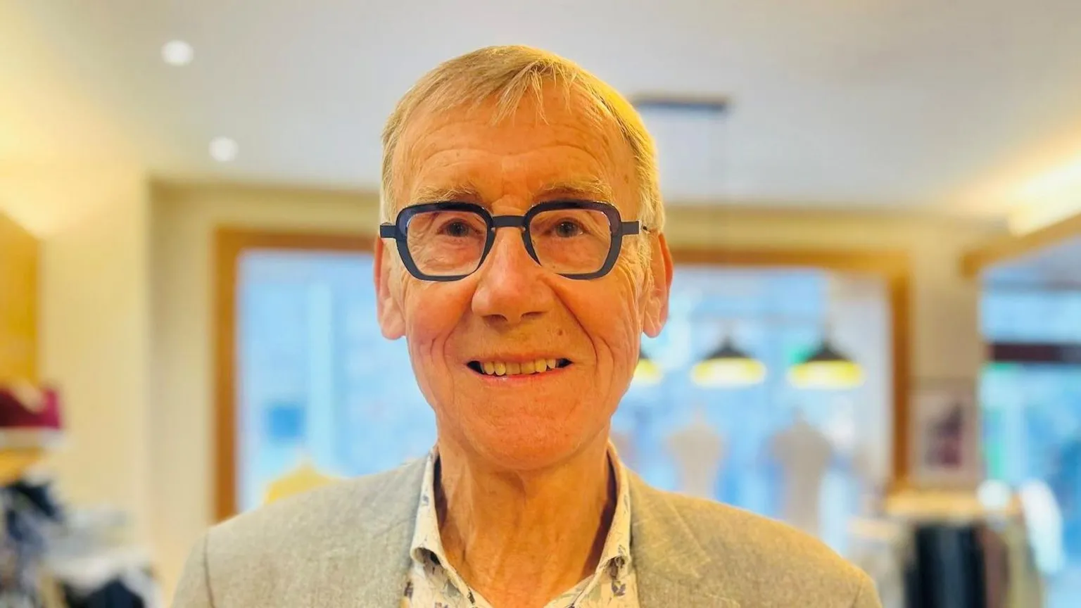 Norman Esslemont, is wearing dark-rimmed glasses, a light-coloured suit jacket and a bright patterned shirt. He is inside a clothes shop.