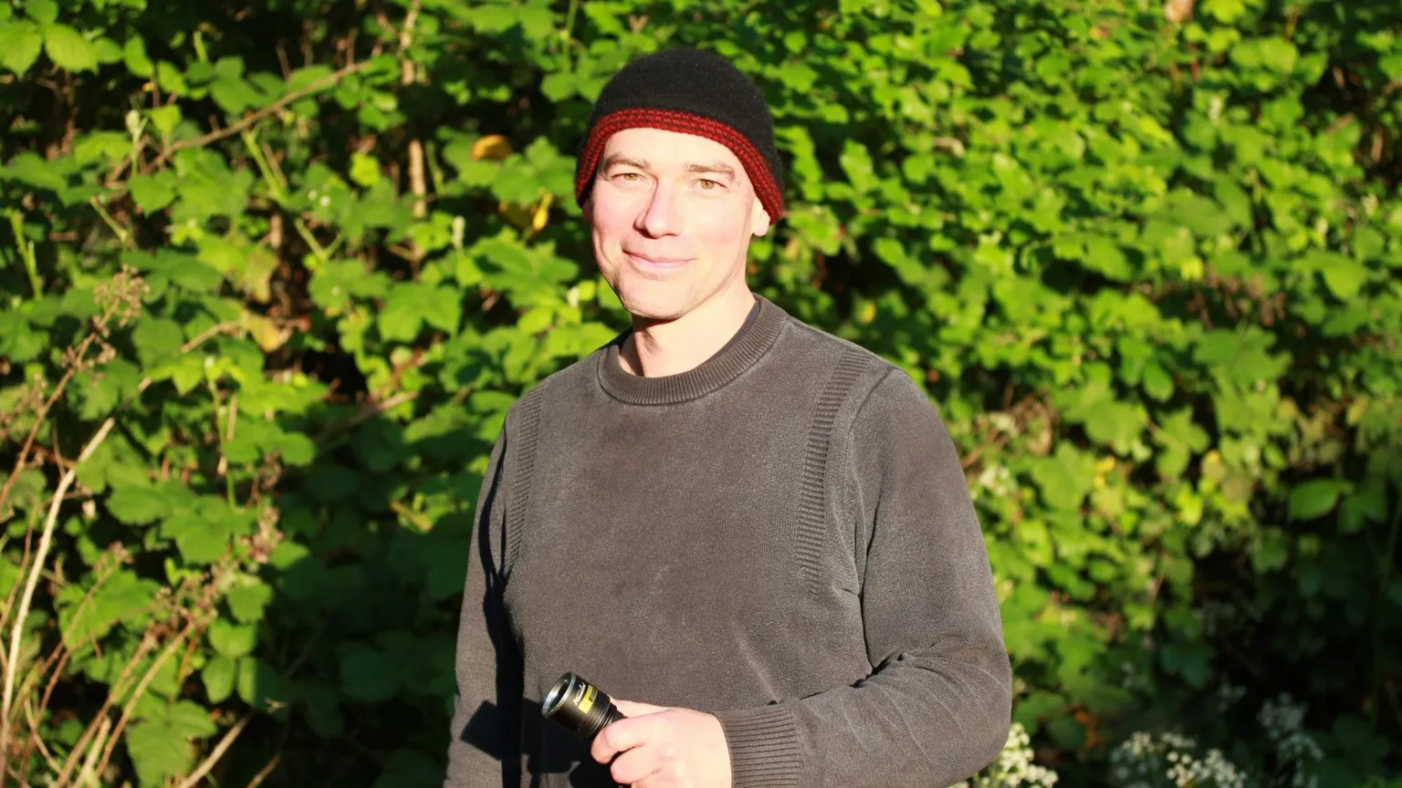 Pete Hughes A man, wearing a dark grey jumper and black and red beanie, is holding a torch and standing in a garden.