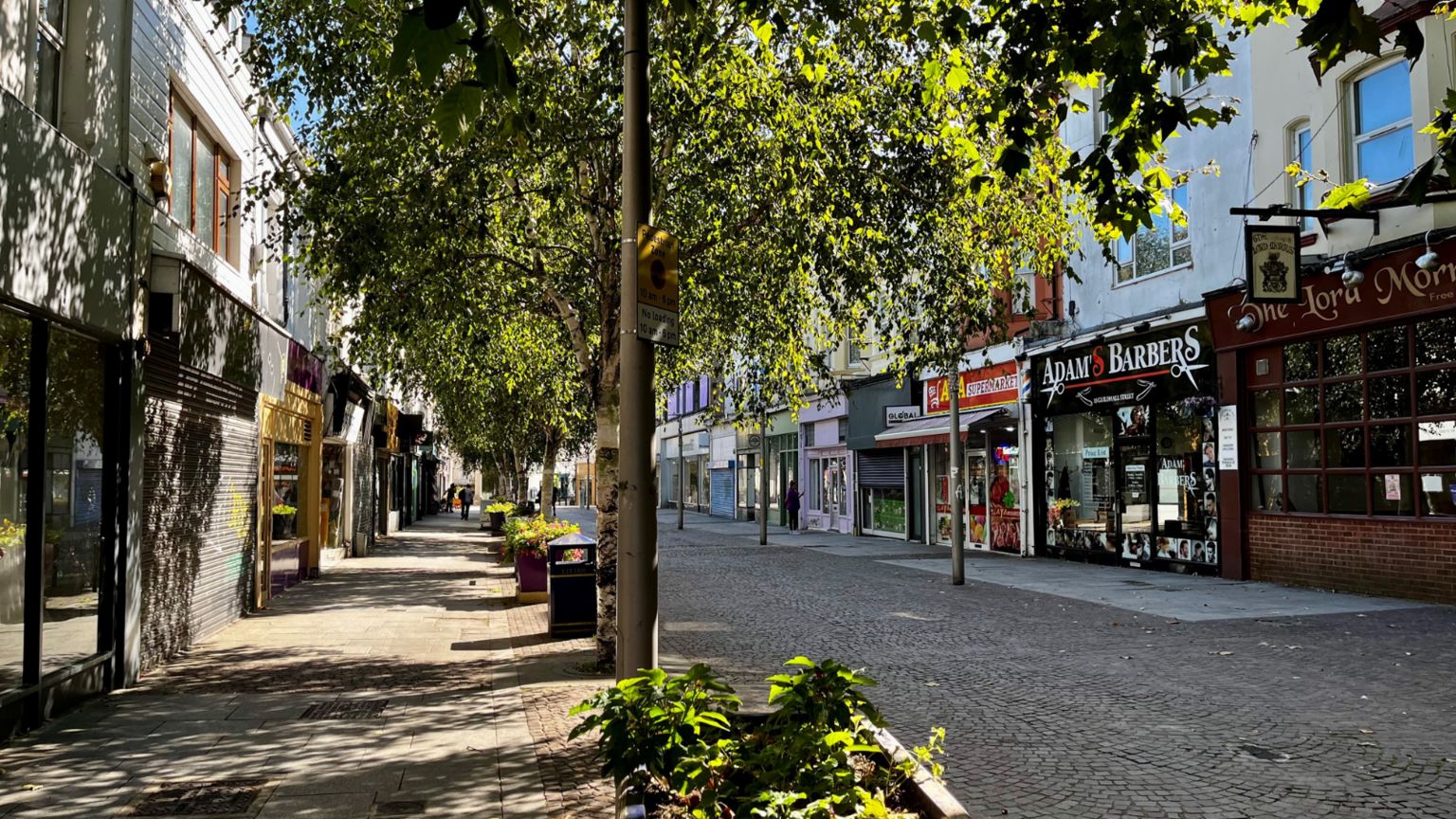 A sunny high street with colourful shops and a line of leafy trees running through it. 