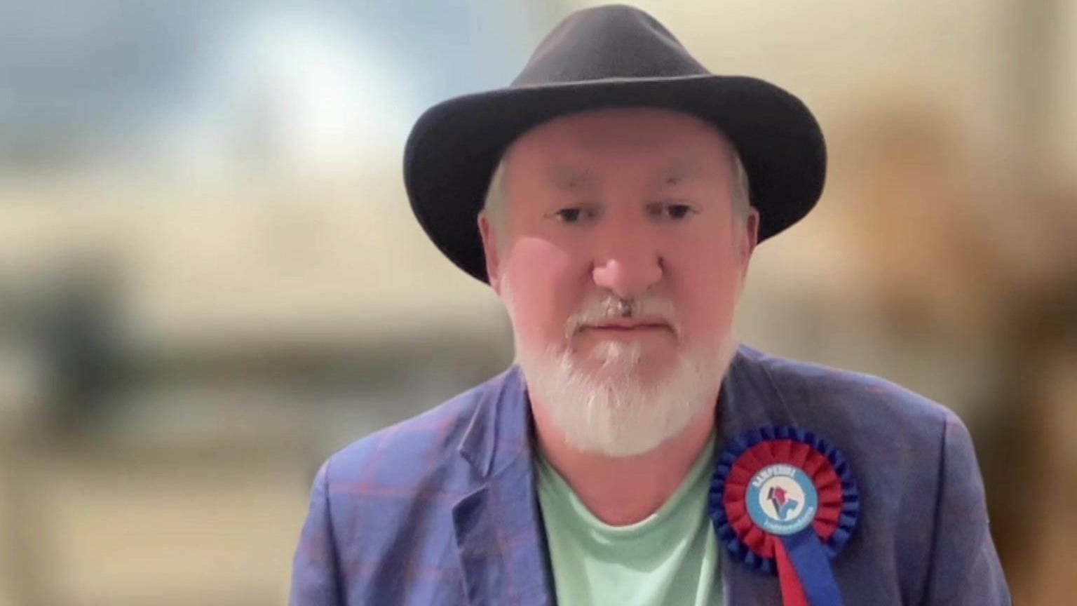 Alan Stone wearing a dark trilby hat and a blue blazer with a red and blue rosette on the lapel