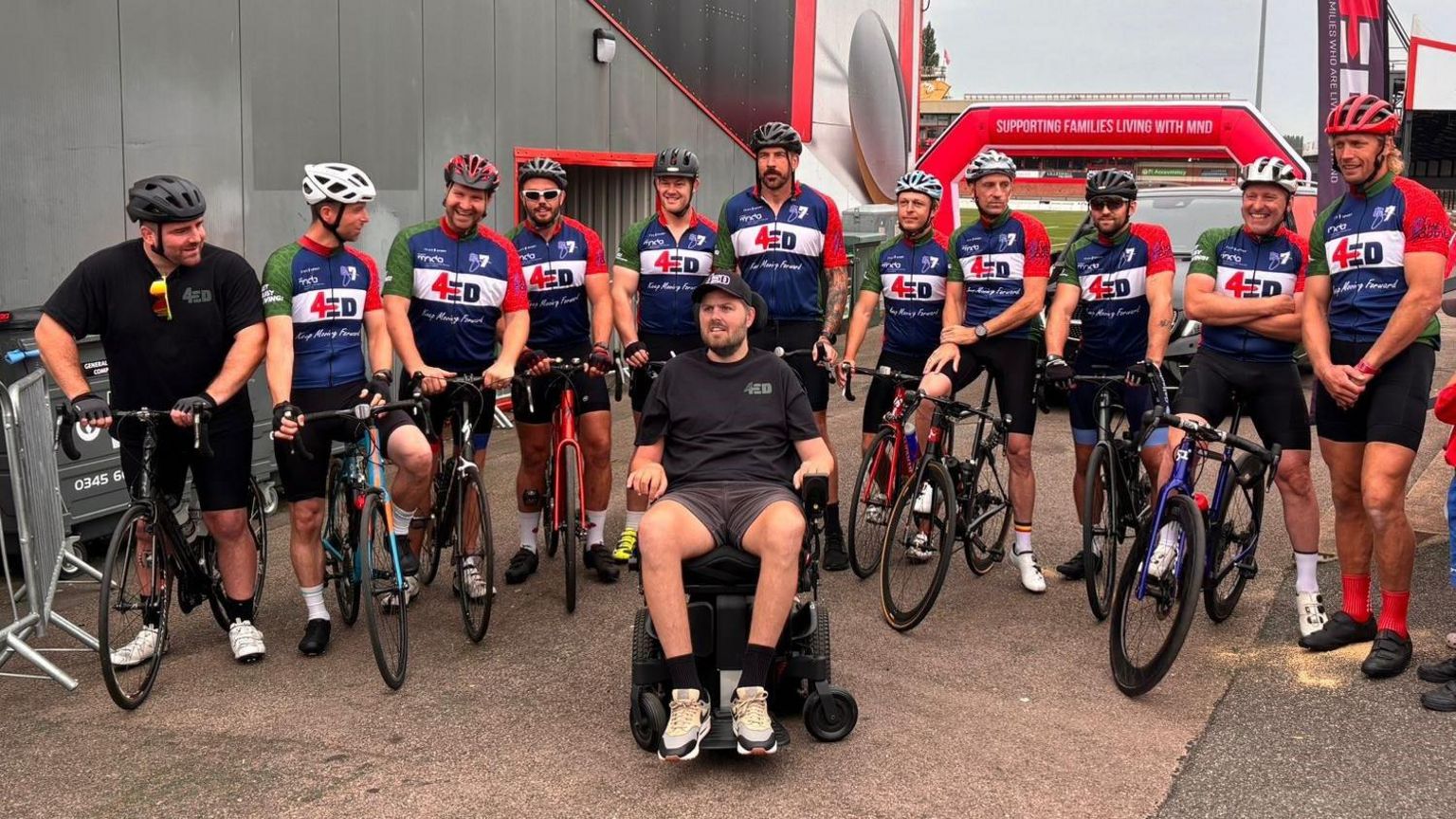 Eleven people sitting on their bikes in a line, with their legs either side. They are all wearing blue, red and white cycling shirts that say '4Ed' on them. Ed Slater is sitting in his wheelchair just in front of them 