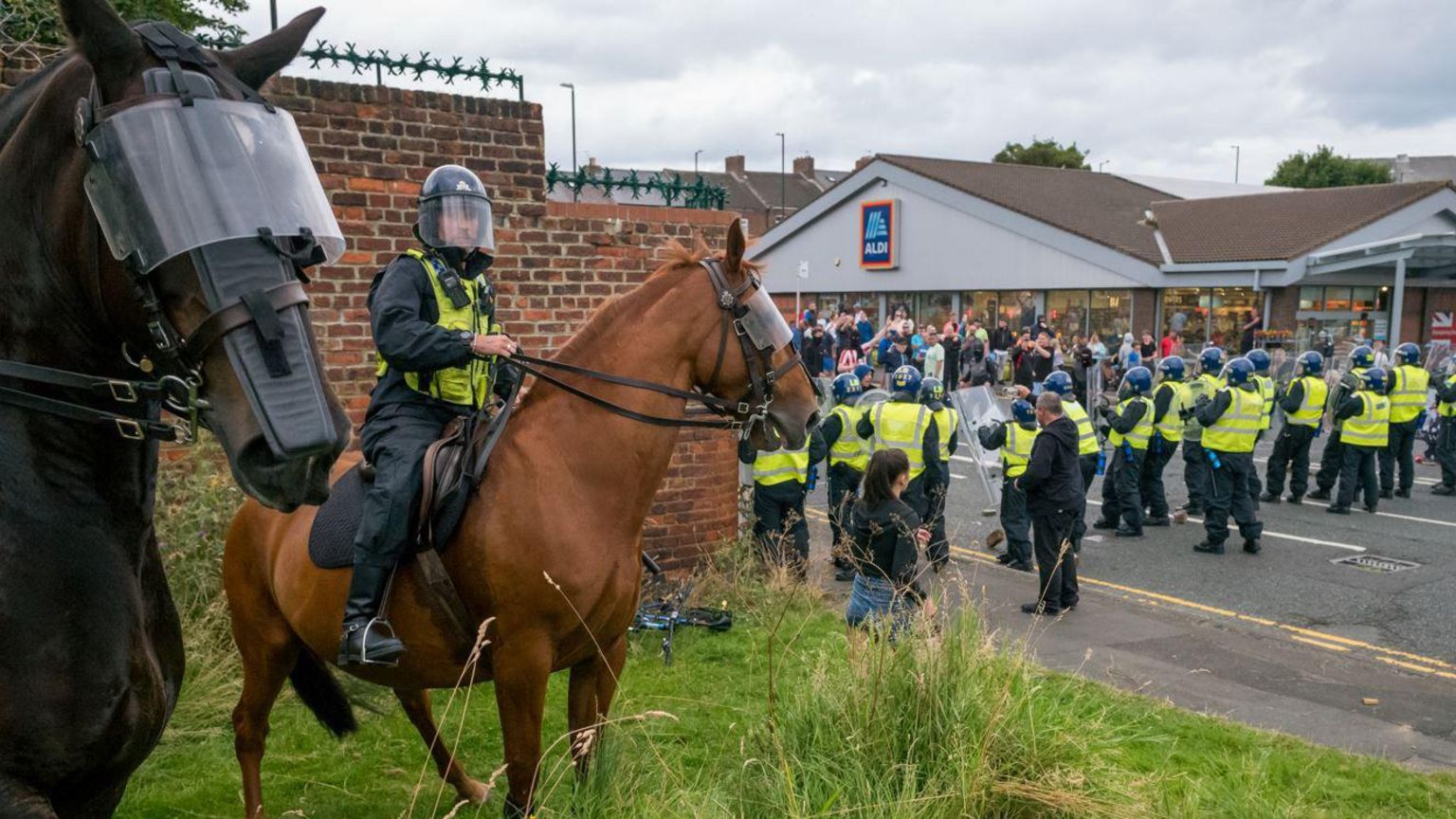 A line of police in riot gear facing rioters outside an Aldi in Sunderland. Police officers on horseback stand in the foreground.