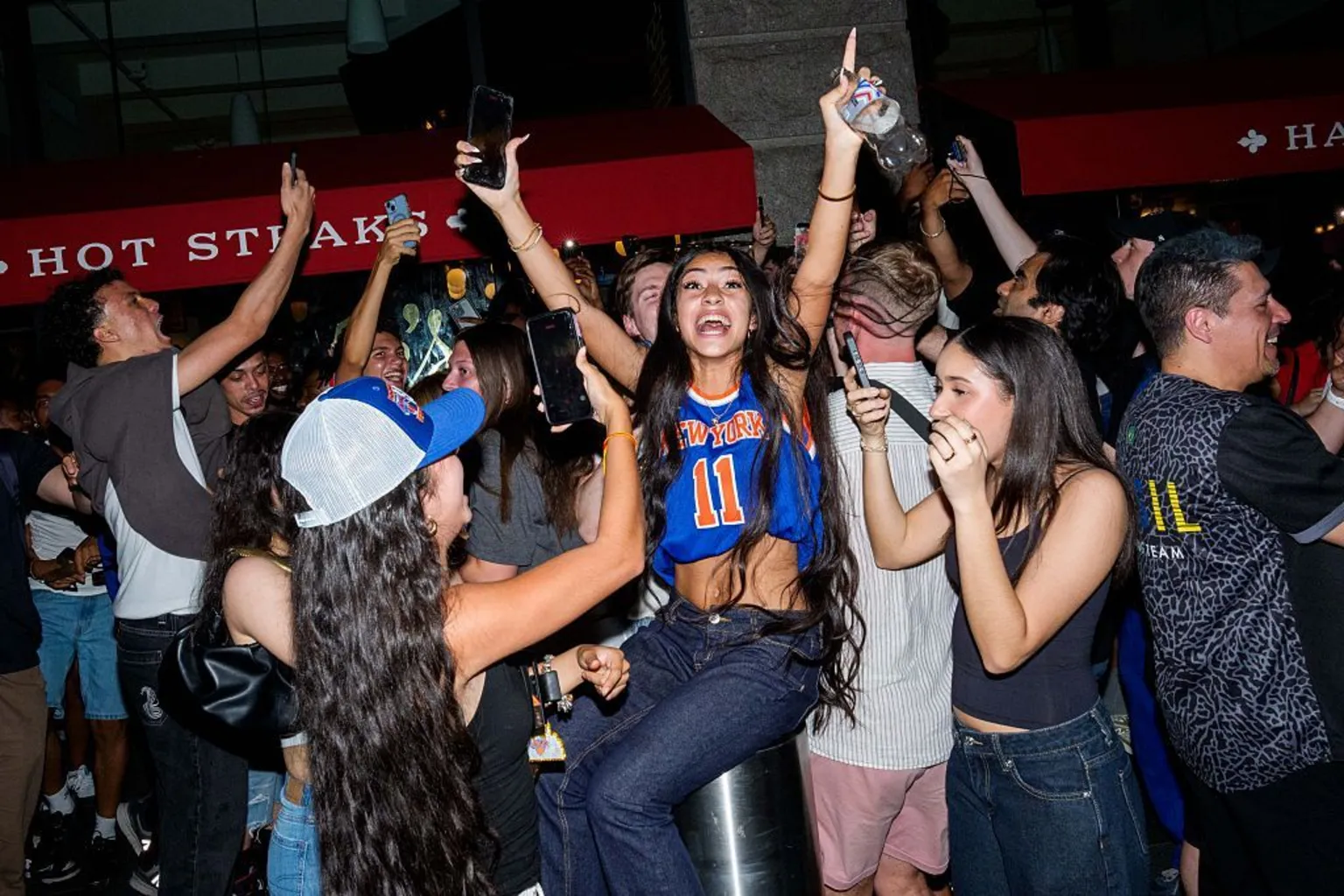 Getty Images A young woman wearing a Knicks jersey raises her hands in the air in celebration, surrounded by other young people.