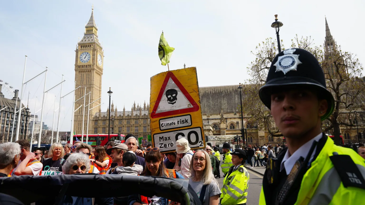 The image shows a climate protest in Westminster with Big Ben visible in the background. A prominent yellow sign displays a skull symbol warning 