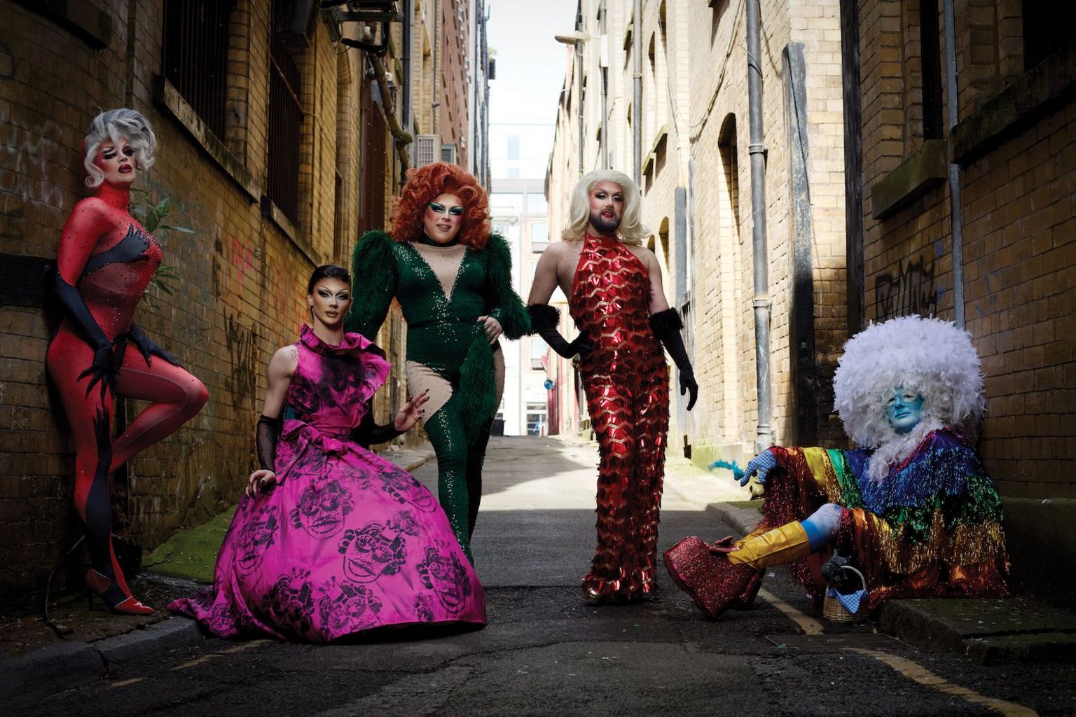 Five drag queens pose in an alley way. One in a red jumpsuit covered in handprints, one in a pink floral gown, another in a green sparkly jumpsuit, a fourth in a red dress decorated in fish scales and the fifth in a rainbow tasseled outfit with a white feathered afro wig.