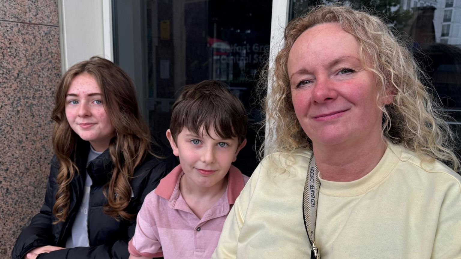 Olivia, Daniel and Rita - a young girl with light brown hair wears a black puff jacket, a young boy wears a pink polo shirt and a woman with curly blonde hair wears a yellow crewneck jumper