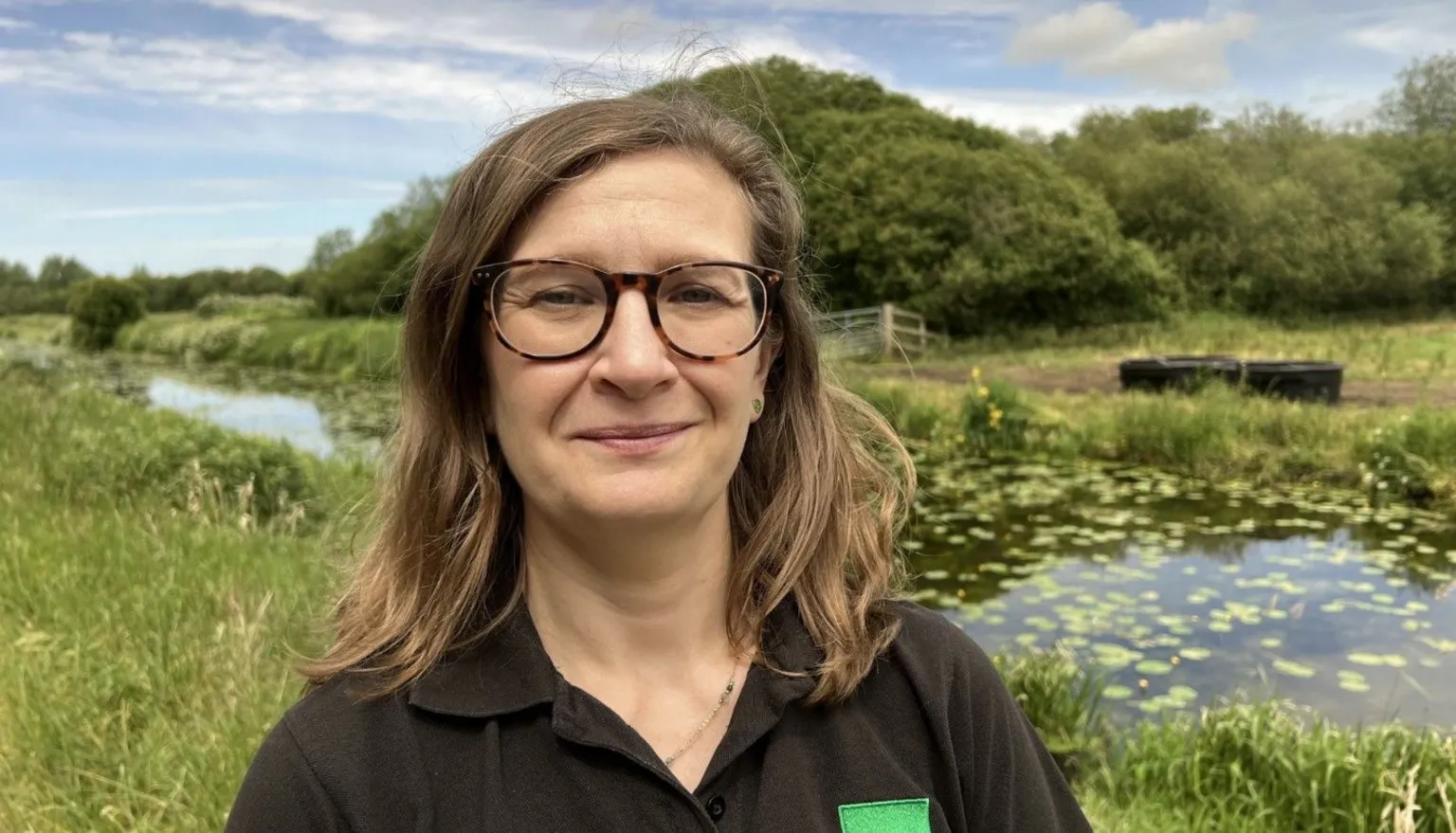 Claire Newill is standing in a field in front of a river. She is wearing a black shirt and glasses and has medium length brown hair.