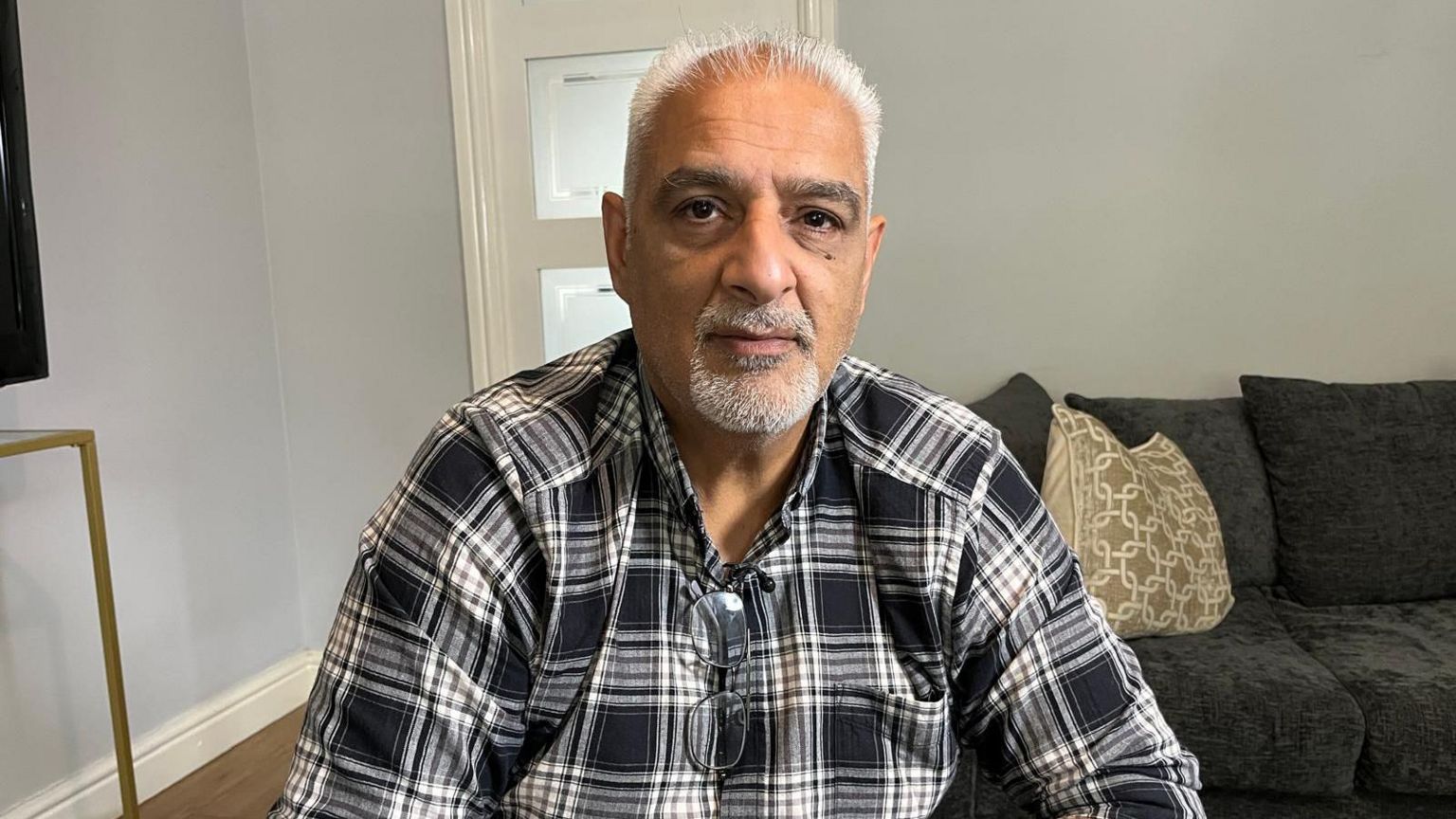 Tariq Jahan at his home. He is wearing a checked shirt with glasses tucked in the front.