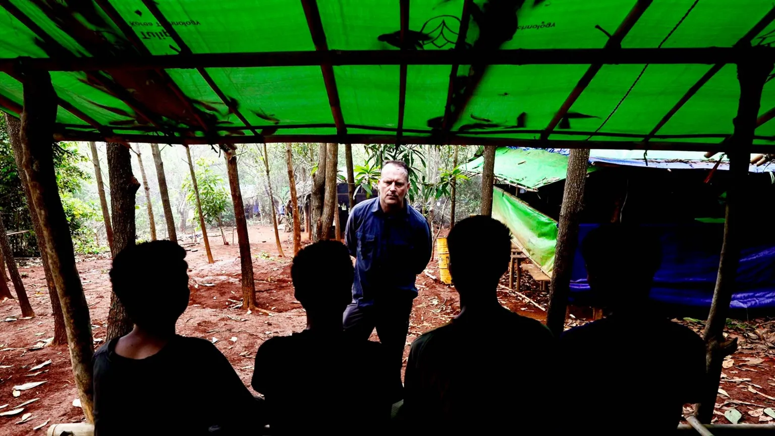 BBC/Darren Conway Four young men stand with their backs to the camera, in a jungle under a tarpaulin roof. BBC correspondent Quentin Sommerville stands facing towards them.