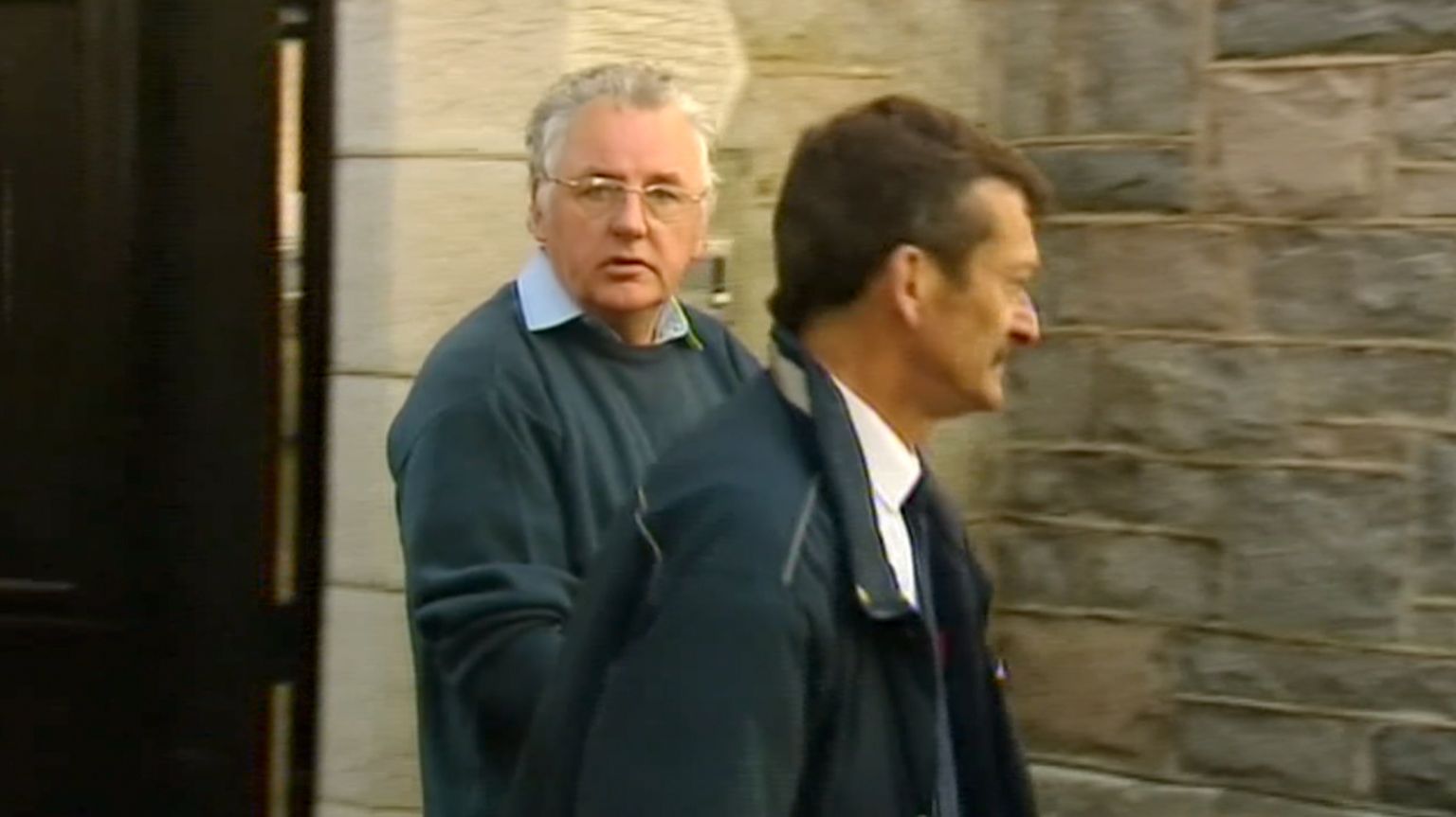 Noel Thomas wearing a blue sweater and blue shirt is led away by a security officer at the old Caernarfon Crown Court