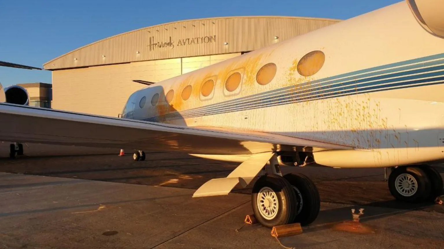 A white private jet is parked on an airport tarmac with orange wheel chocks. The aircraft shows visible orange paint damage along its fuselage near the passenger windows. 