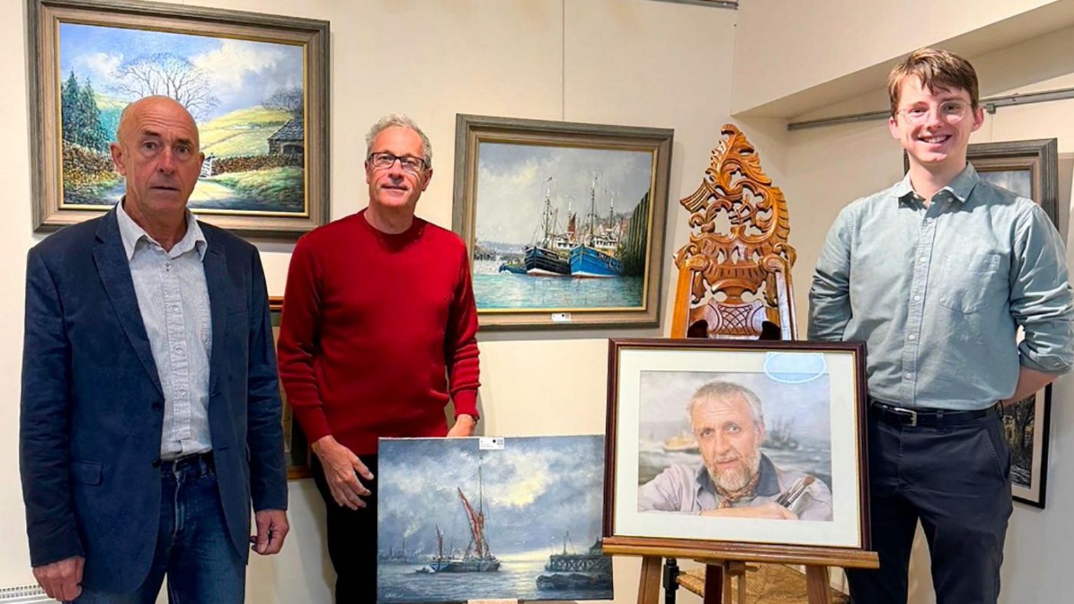 Three men: one wearing a blue suit, another with a red jumper and black-framed glasses and a younger man smiling and wearing a light-blue shirt, dark blue trousers and metal-framed circular glasses. They are standing among paintings