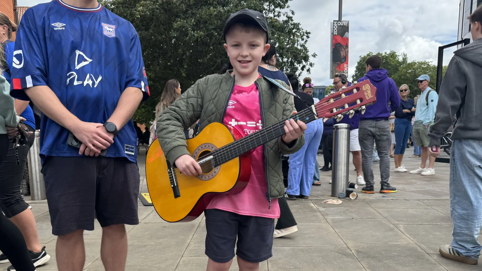 Zoie O'Brien/BBC A boy holds an acoustic guitar. He wears a dark cap, a green coat, a pink Ipswich Town shirt and shorts.