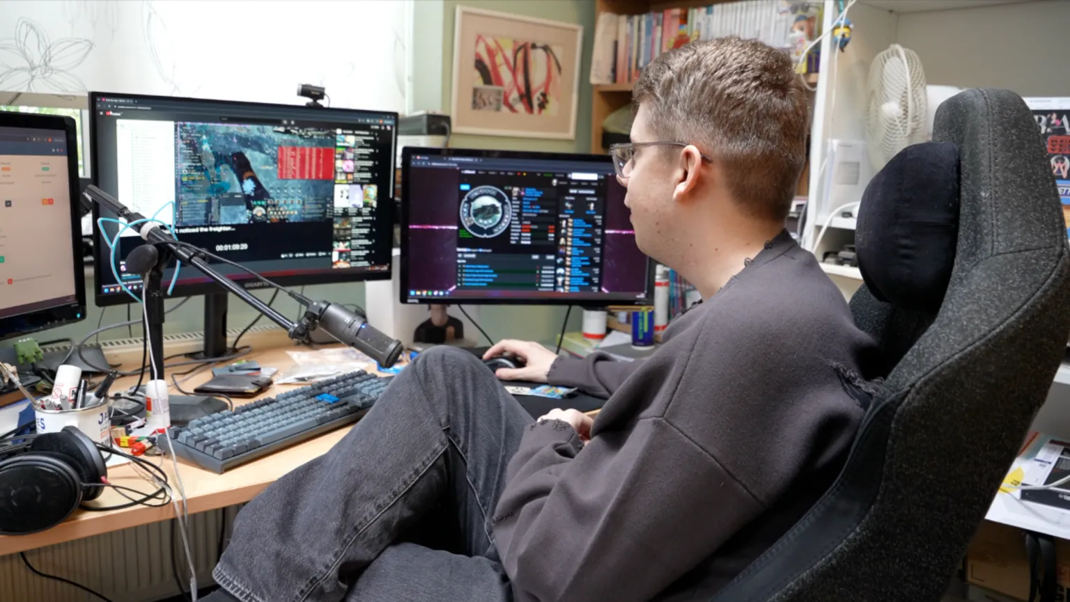 James Cunningham sits at a multi-monitor desk setup playing EVE Online, with the game interface open on one screen and other windows visible on adjacent displays. A keyboard, microphone, and headphones are arranged across the cluttered workspace.