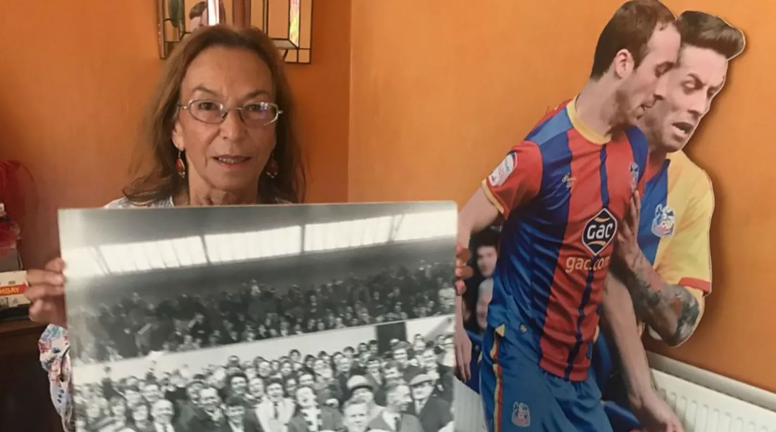 Hy Money holding a black and white picture of a crowded football stadium. Behind her is a life-sized cutout of two Crystal Palace football players in action.