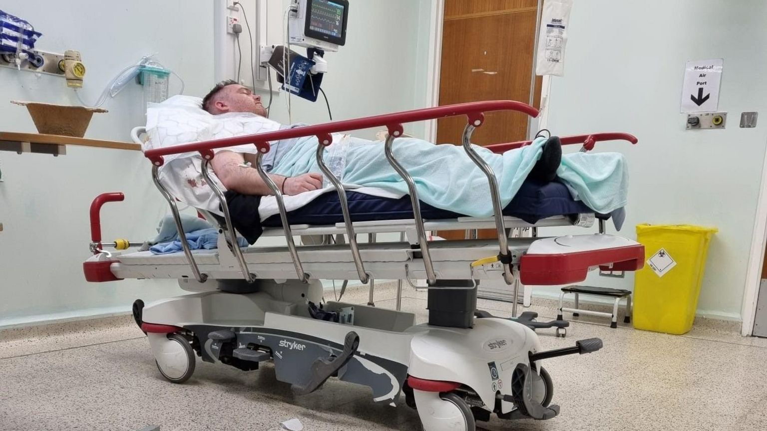 PC Aspinall lies in a hospital bed covered with a blanket. His eyes are closed and he appears to be hooked up to machines. 