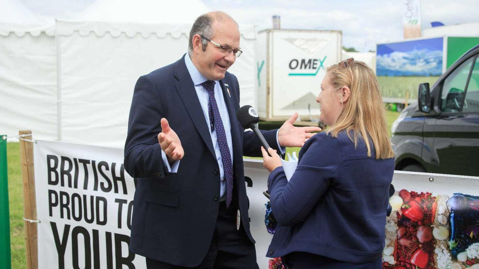 Milly Fyfe has long blond hair and is wearing a blue jacket and holding a microphone. She is interviewing Tom Bradshaw who has short dark hair and is wearing a dark suit and is standing outdoors next to a large sign.