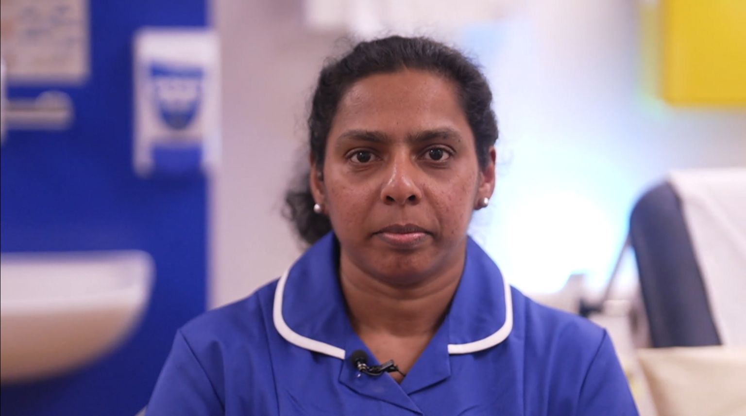 Looking directly at the camera is Tincy Jose wearing her blue nursing uniform which has a white lining around the collar. She has dark hair, brown eyes and white stud earrings. She is inside the Queen Elizabeth Hospital in King's Lynn and behind her there is a hospital bed and sink. 

